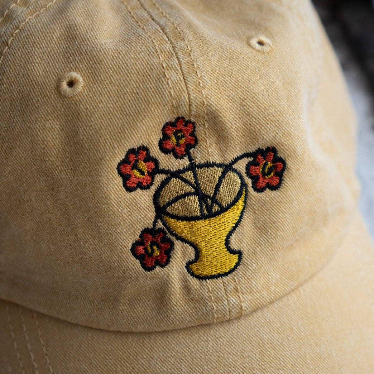 Beige cap with embroidered design of a yellow vase with flowers on a plain background