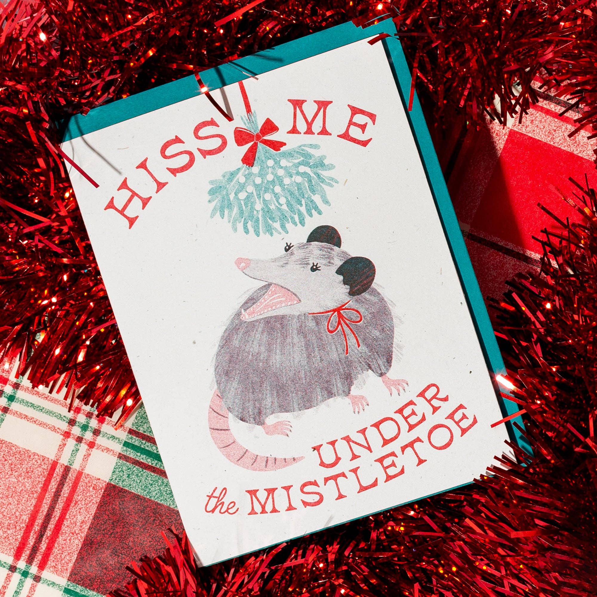Christmas card with a opossum illustration and text 'Kiss me under the mistletoe' on a red tinsel background.