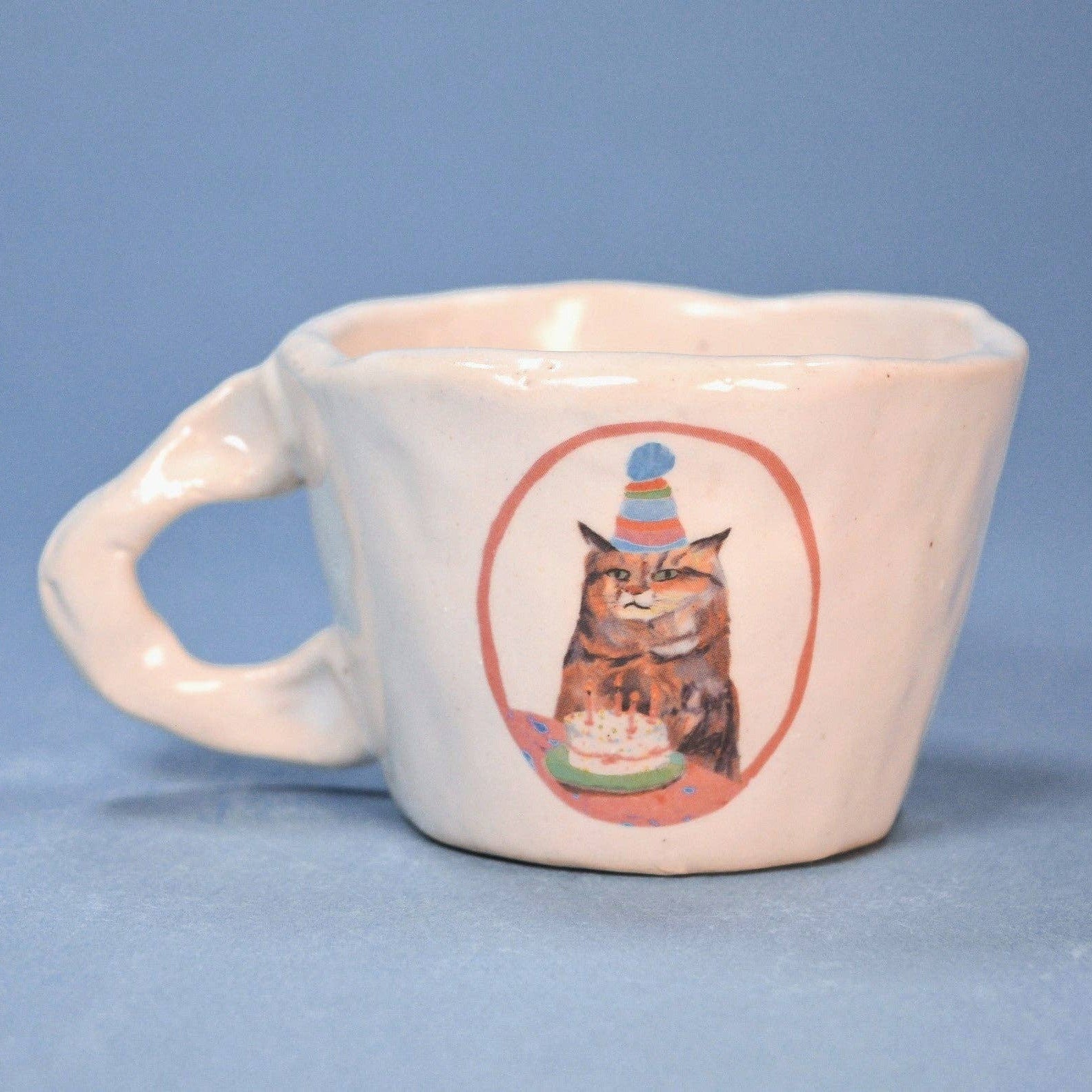 White ceramic mug with a cat wearing a party hat and birthday cake design on a blue background