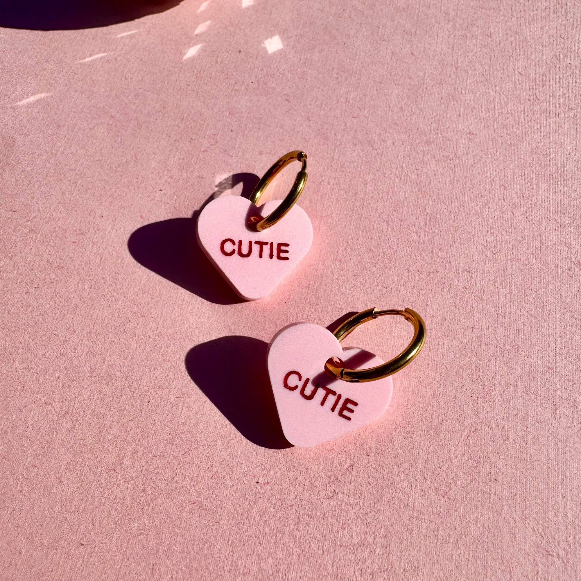 Heart-shaped 'CUTIE' keychains on a pink surface with a disco ball in the background.