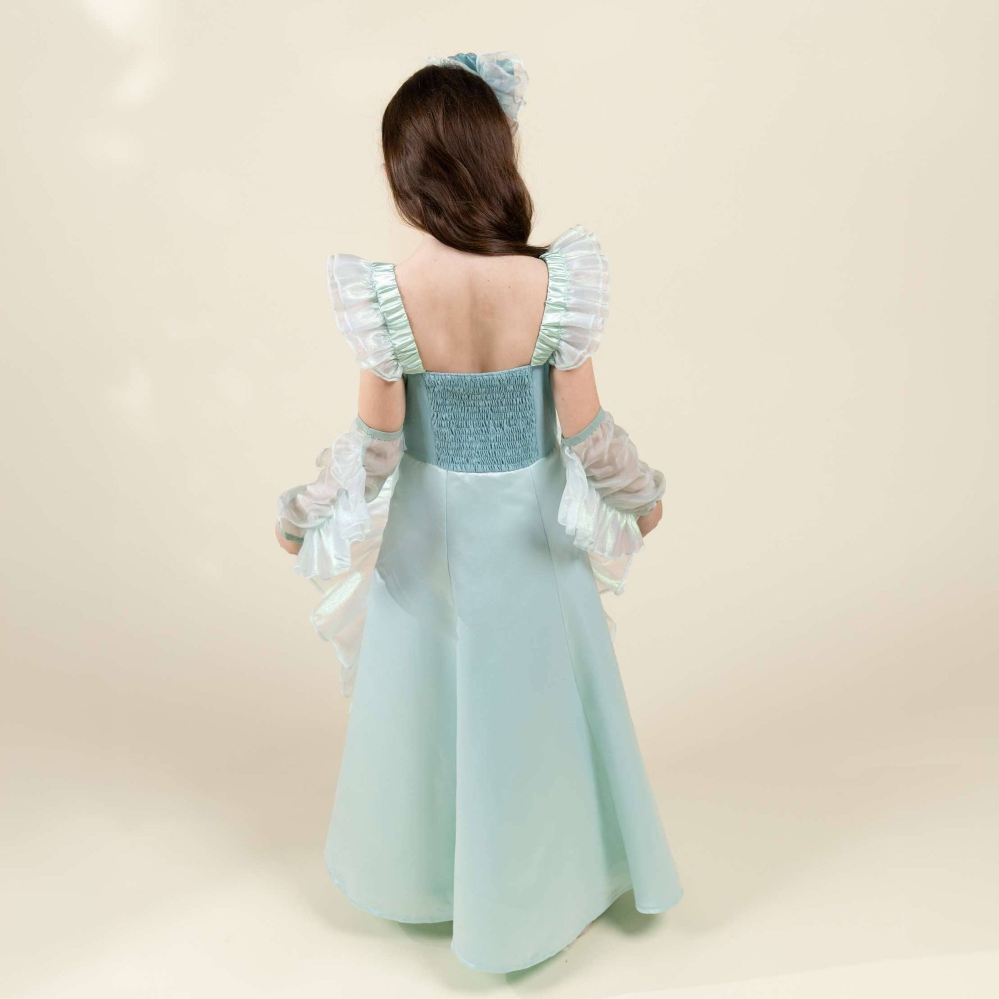 A young girl viewed from behind wearing a light blue princess-style dress with ruffled sleeves, sheer long gloves, and a matching hair bow, photographed against a soft cream studio background.
