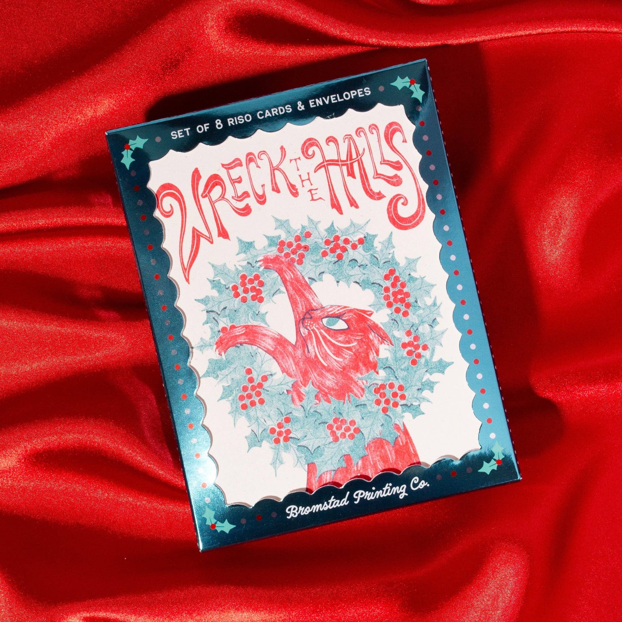 A boxed card set titled "Wreck of the Titanic" by Bromsted Printing Co, shown on draped red satin fabric. The box has a white center with red script lettering, a red bird illustration surrounded by blue holly leaves, and a blue scalloped border.