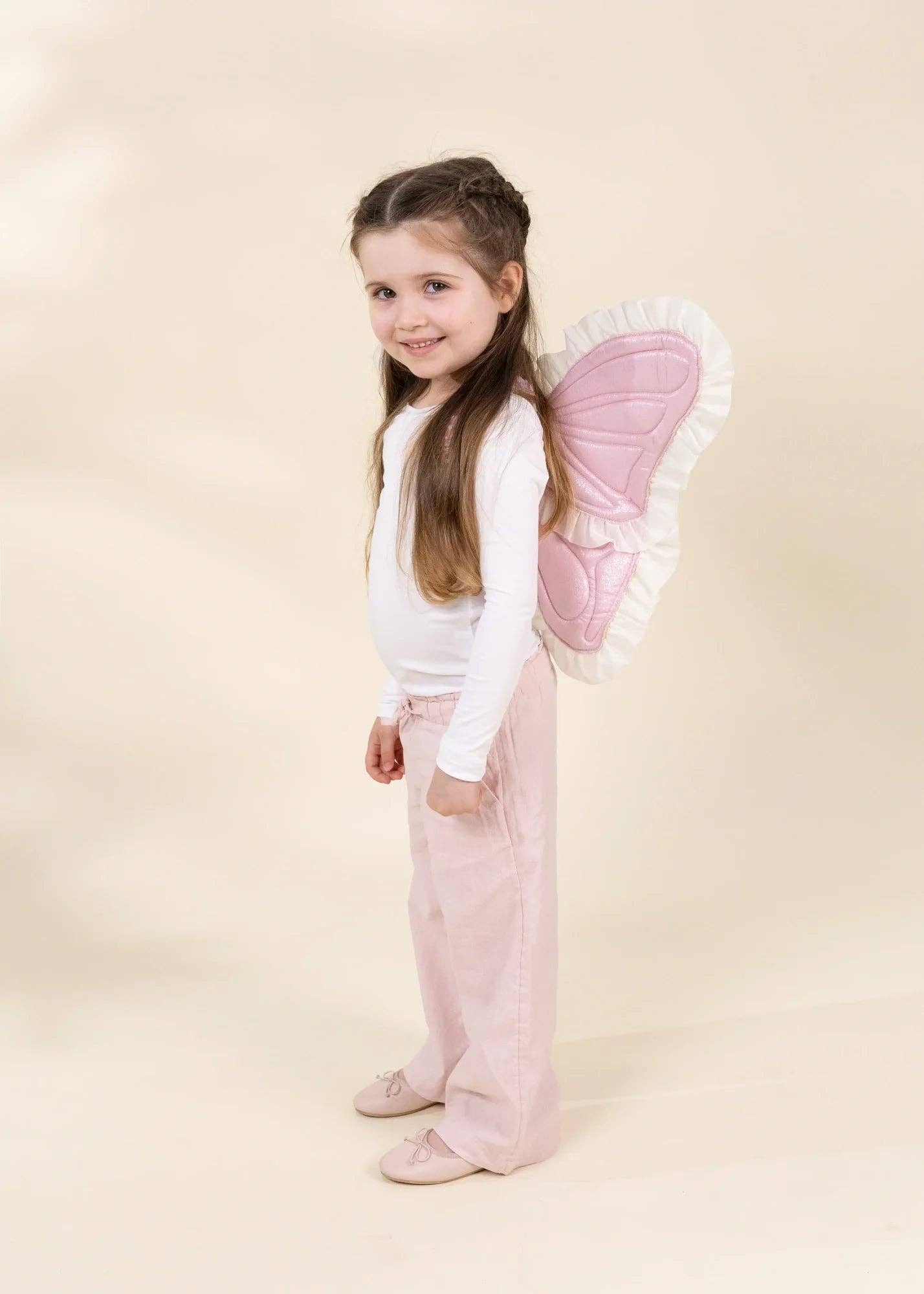 A young girl with long brown hair stands in a studio wearing a white long-sleeve top, light pink pants, and matching pink shoes, with a pink and white quilted fairy wing accessory on her back. The background is a solid, warm cream color.