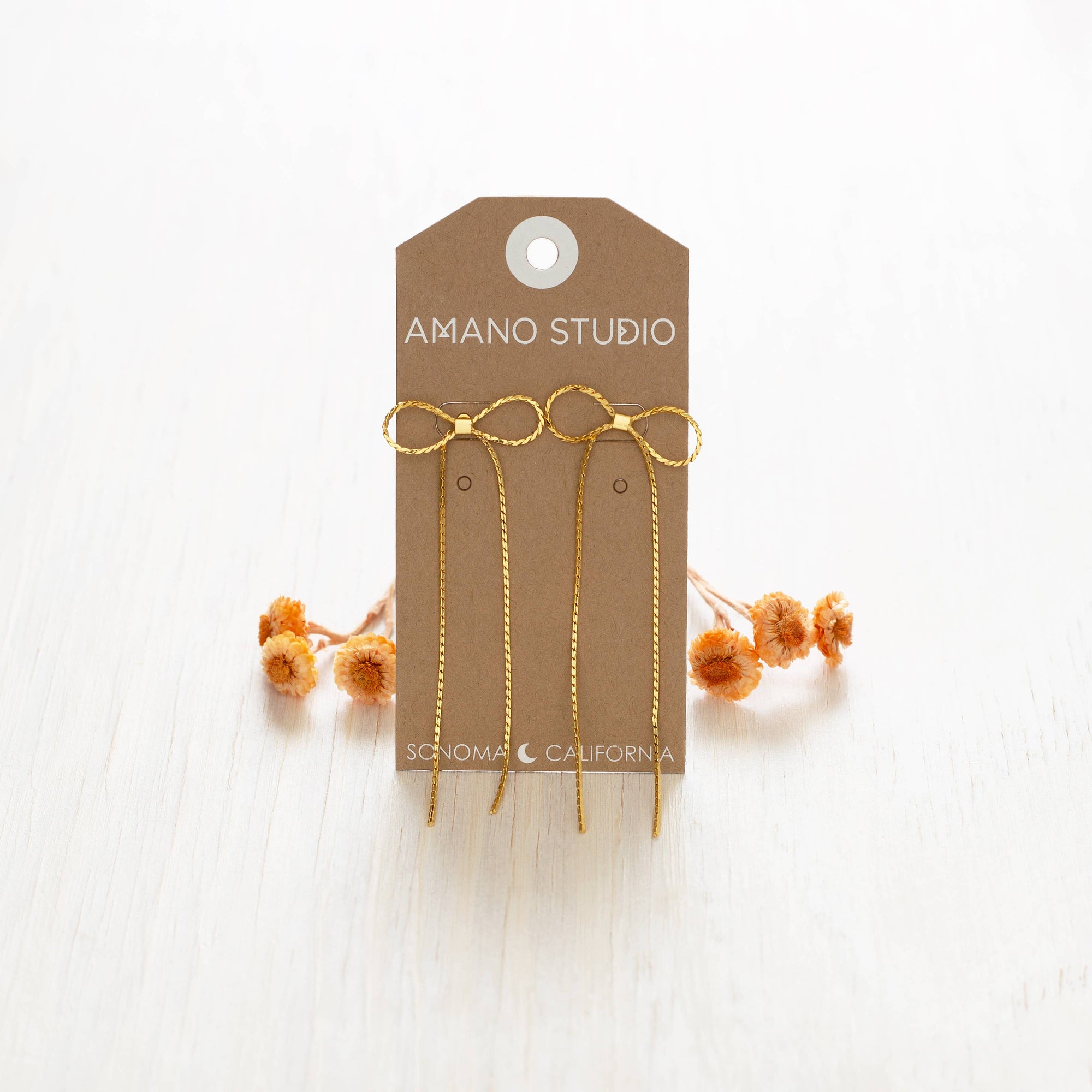 Gold earrings on a 'Amano Studio' branded card with dried flowers on a white background