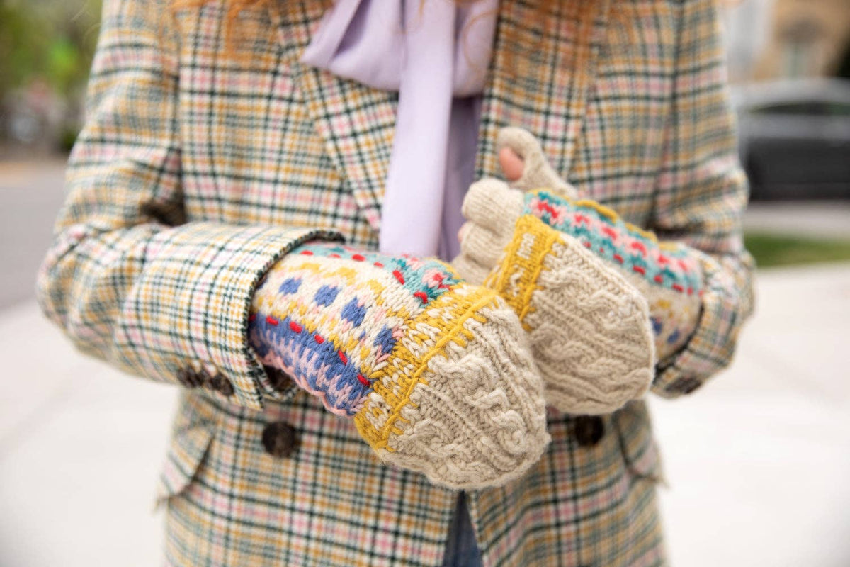 A person wearing a beige plaid coat and a light purple scarf holds up a pair of cream cable-knit winter mittens with colorful Fair Isle patterns in blue, pink, and yellow.
