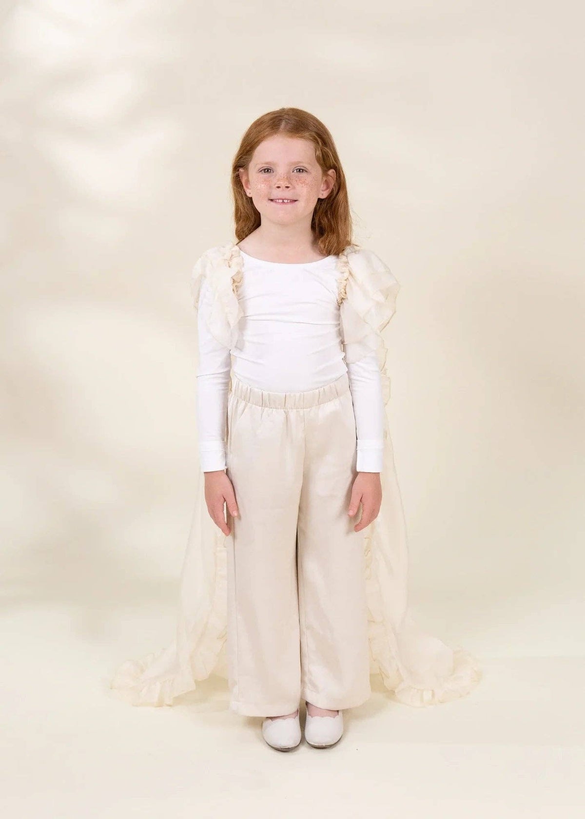 A young girl with red hair stands against a warm cream studio backdrop wearing a white long-sleeve top, cream wide-leg pants, and a matching sheer cream cape with ruffled trim.
