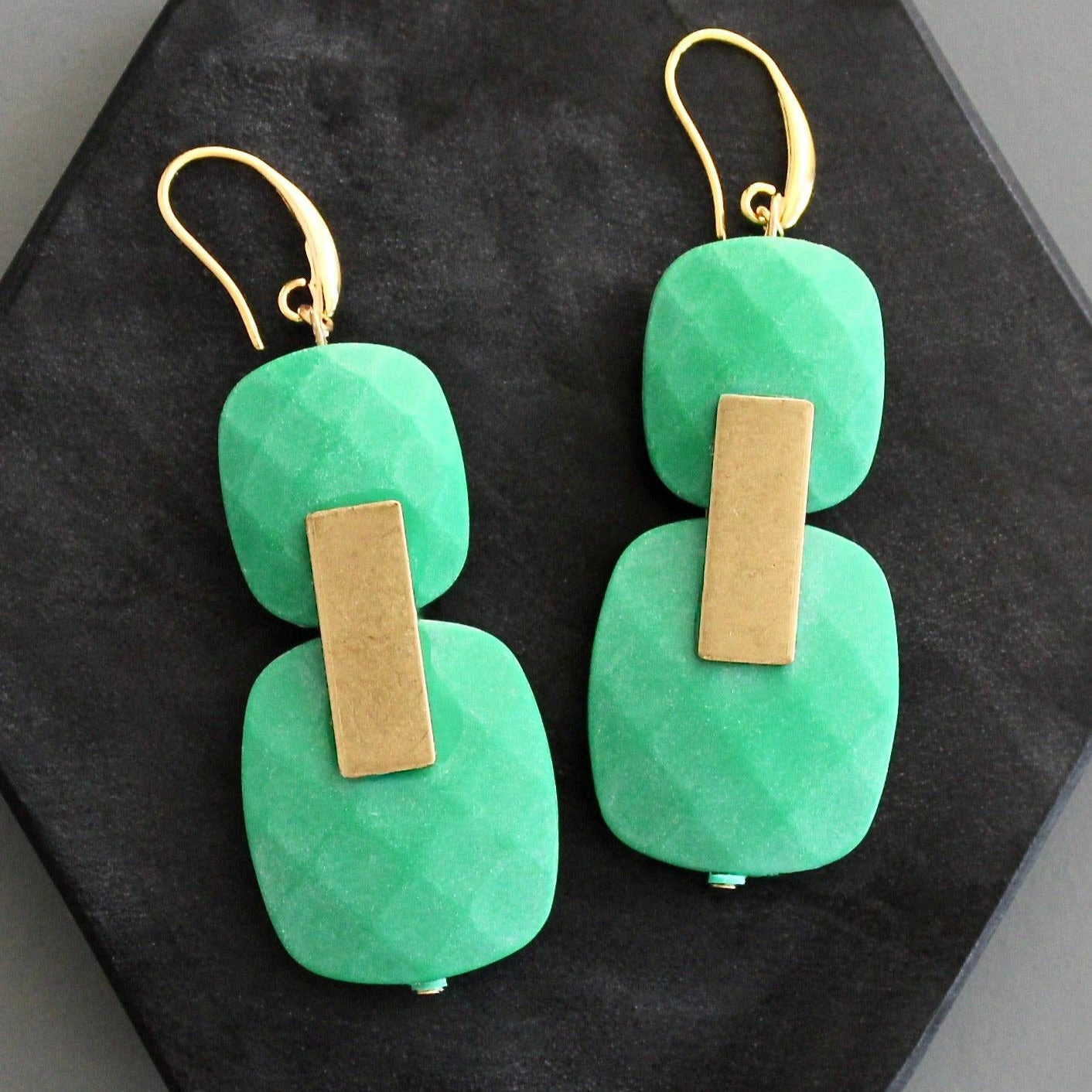 Green earrings with gold accents on a black hexagonal stand against a gray background