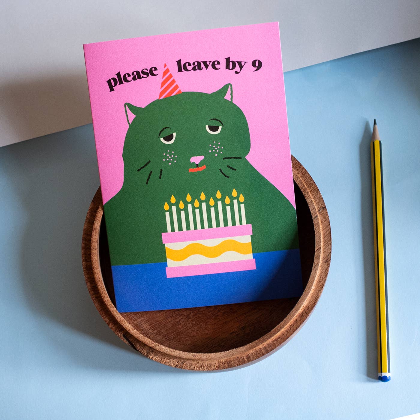 Card with a cat illustration and birthday cake on a pink background, placed in a wooden bowl on a blue surface.
