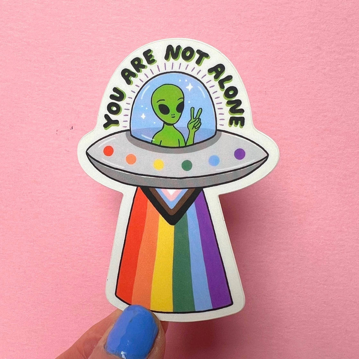 A die-cut sticker featuring a green alien in a UFO with the text 'YOU ARE NOT ALONE' above it, held against a pink background by a hand with blue nail polish.