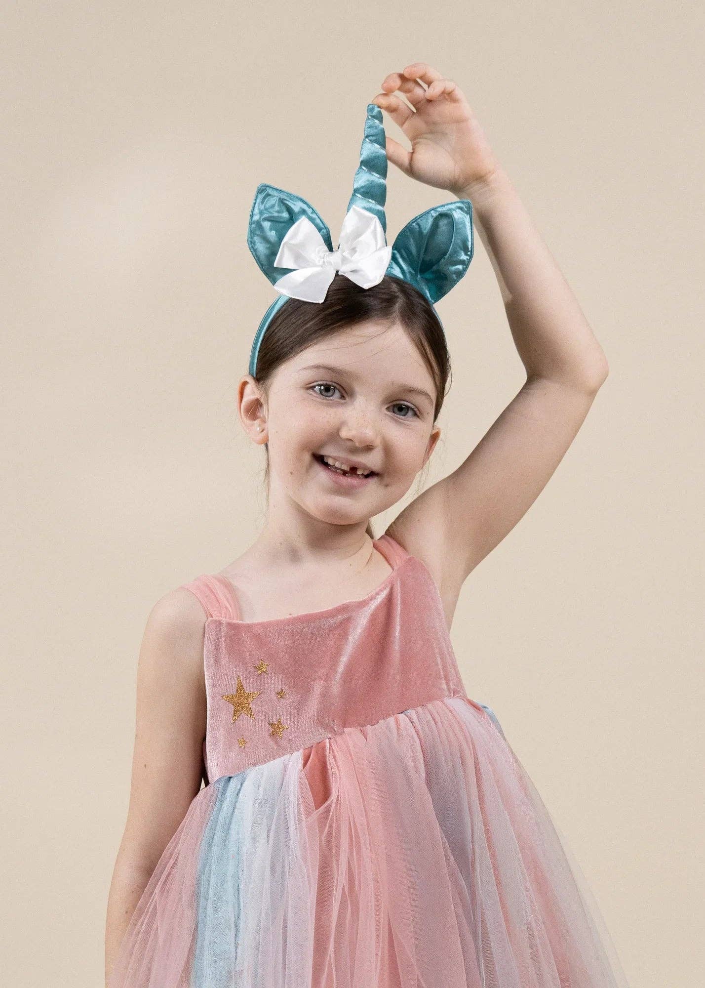 A young girl wearing a pastel unicorn-themed dress and a matching teal and white unicorn headband, posing against a solid beige studio background.