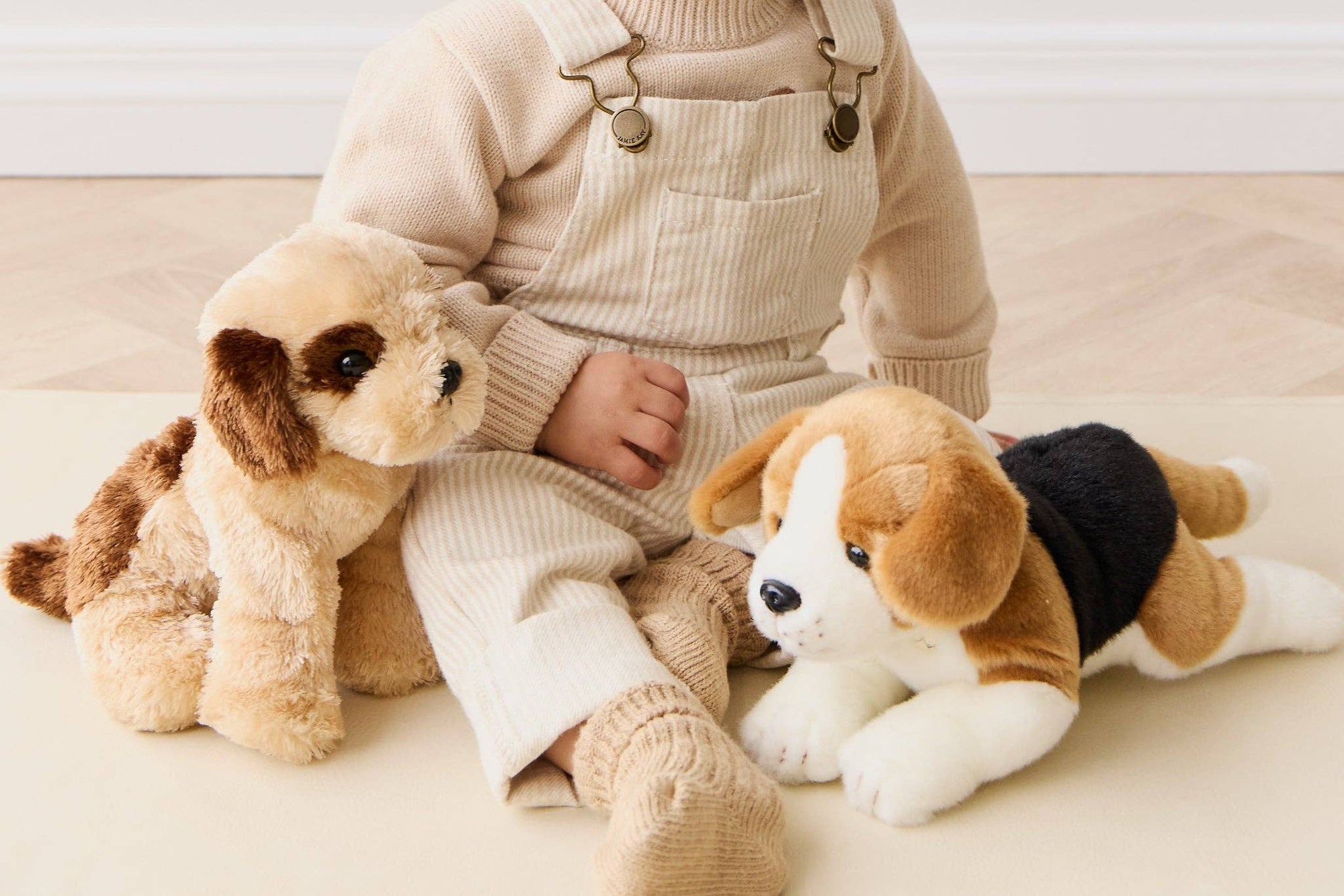 Child in beige outfit with two plush dogs on a light wooden floor.