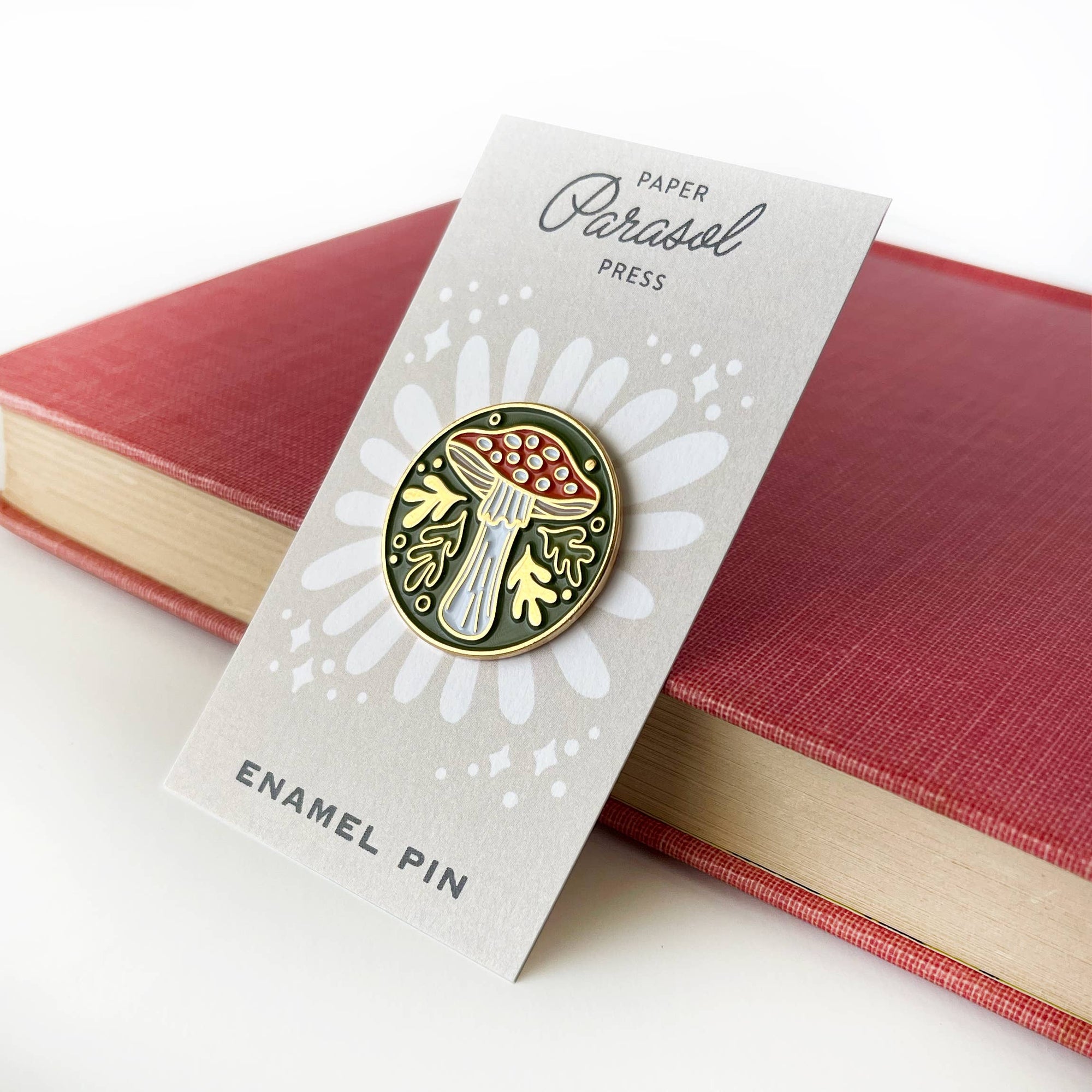 Enamel pin on a 'Paper Parasol Press' card with a red book in the background
