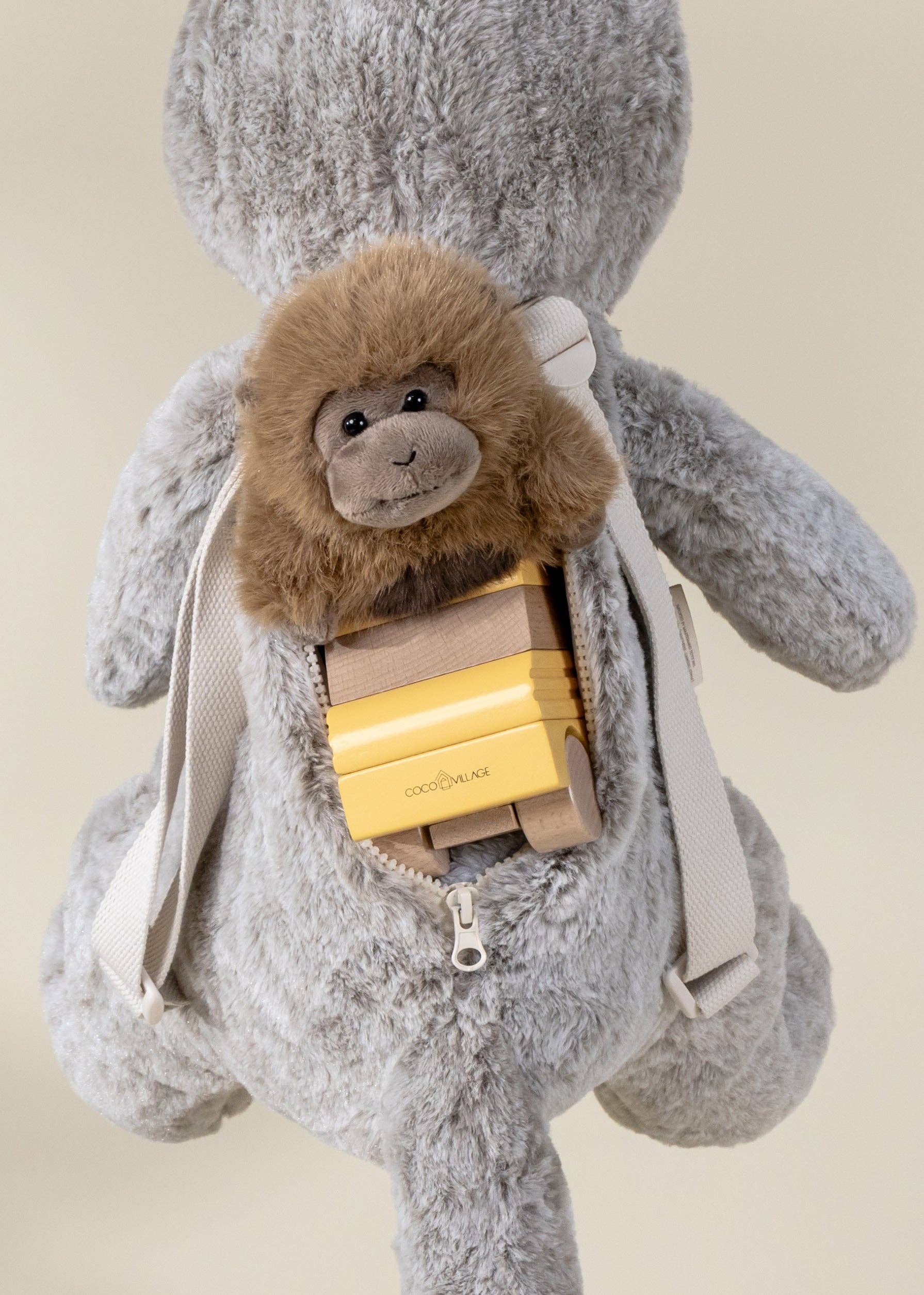 Gray plush bunny with a small monkey plush toy inside a backpack on a beige background