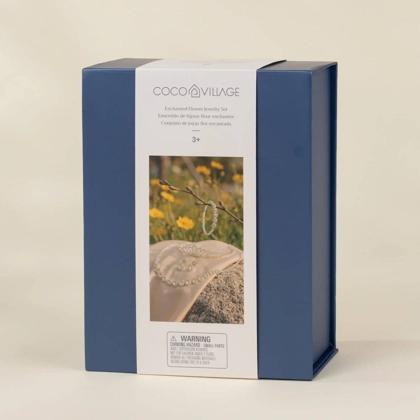 COCO VILAGE brand product box in navy blue and white packaging, featuring a photo of a beaded necklace on a stone with yellow flowers, set against a warm beige background.