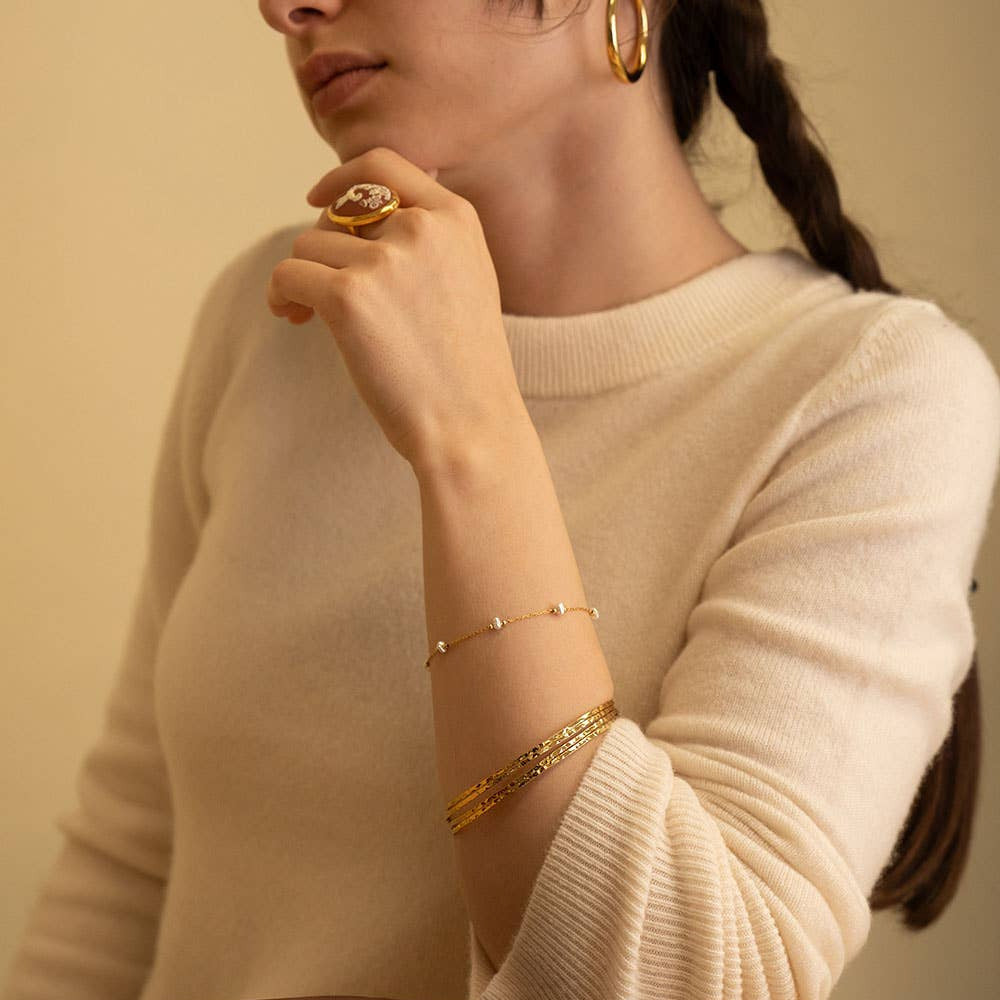 Person wearing gold jewelry including a ring, bracelet, and earrings on a neutral background