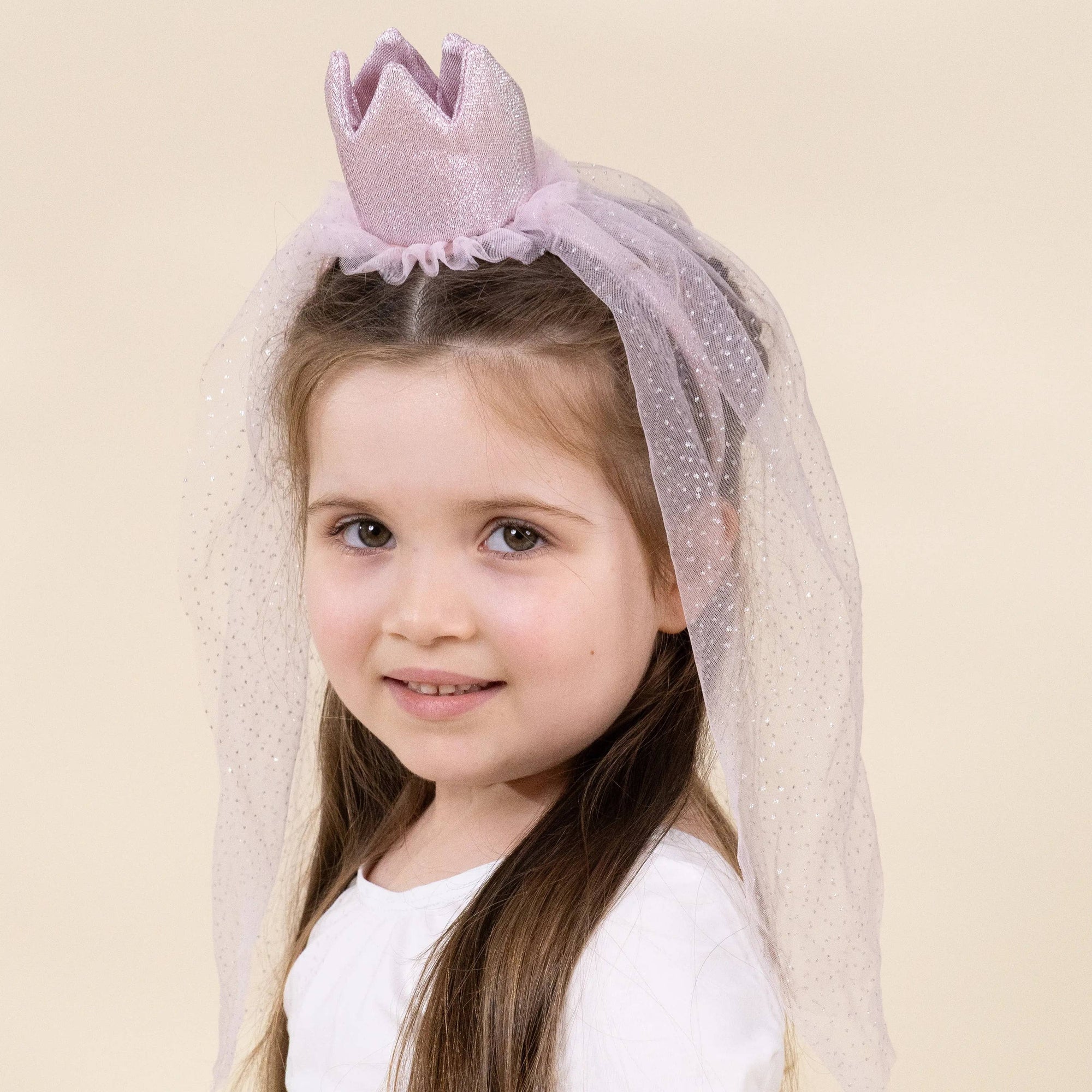 A young girl with long brown hair wearing a light purple glittery tiara and matching sheer tulle veil, paired with a white long-sleeve shirt, against a solid beige studio background.