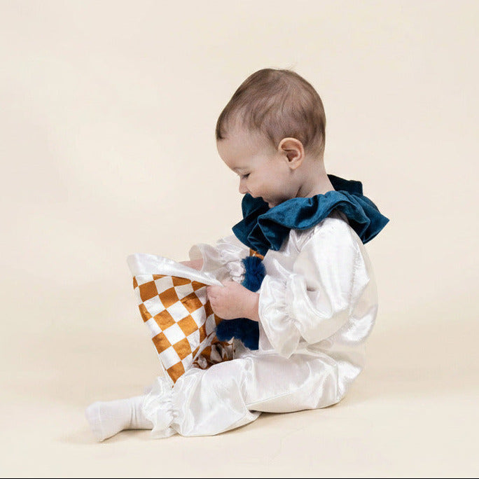 A baby wearing a white satin clown costume with a dark blue ruffled collar sits on a beige studio background, holding a brown and white checkered paper bag.