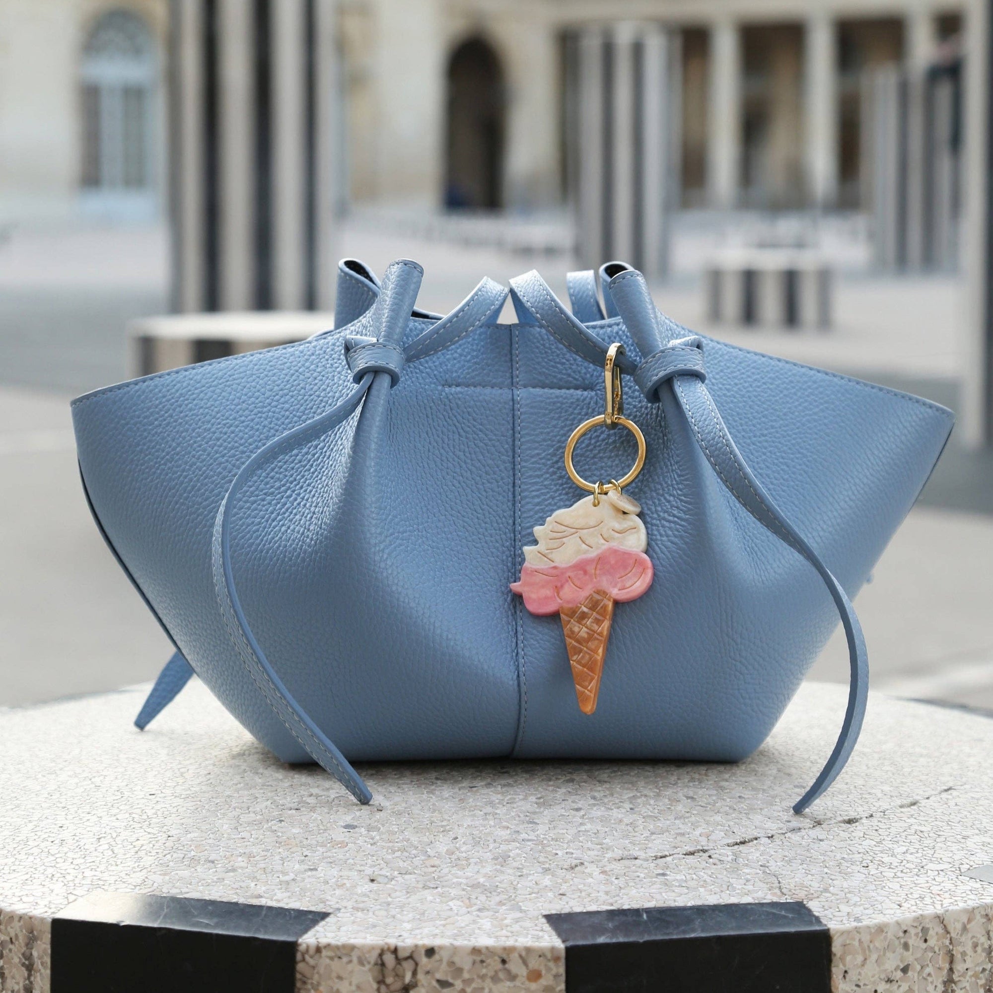 Blue handbag with an ice cream charm on a stone surface, blurred building in the background