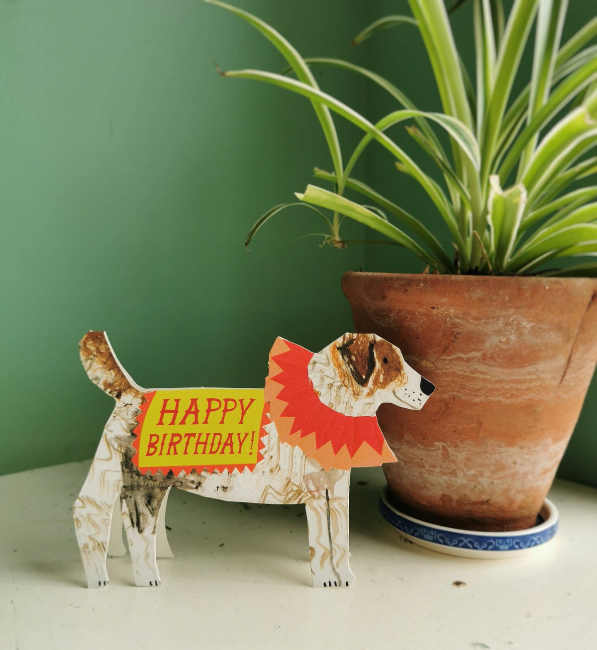 Dog-shaped birthday card with 'Happy Birthday!' message next to a potted plant on a green background