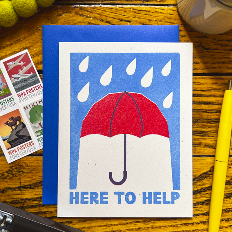 A square art print or card featuring a red umbrella in the rain with the text 'HERE TO HELP' on a white card, placed on a blue backing over a wooden tabletop with nearby pencils and postcards.