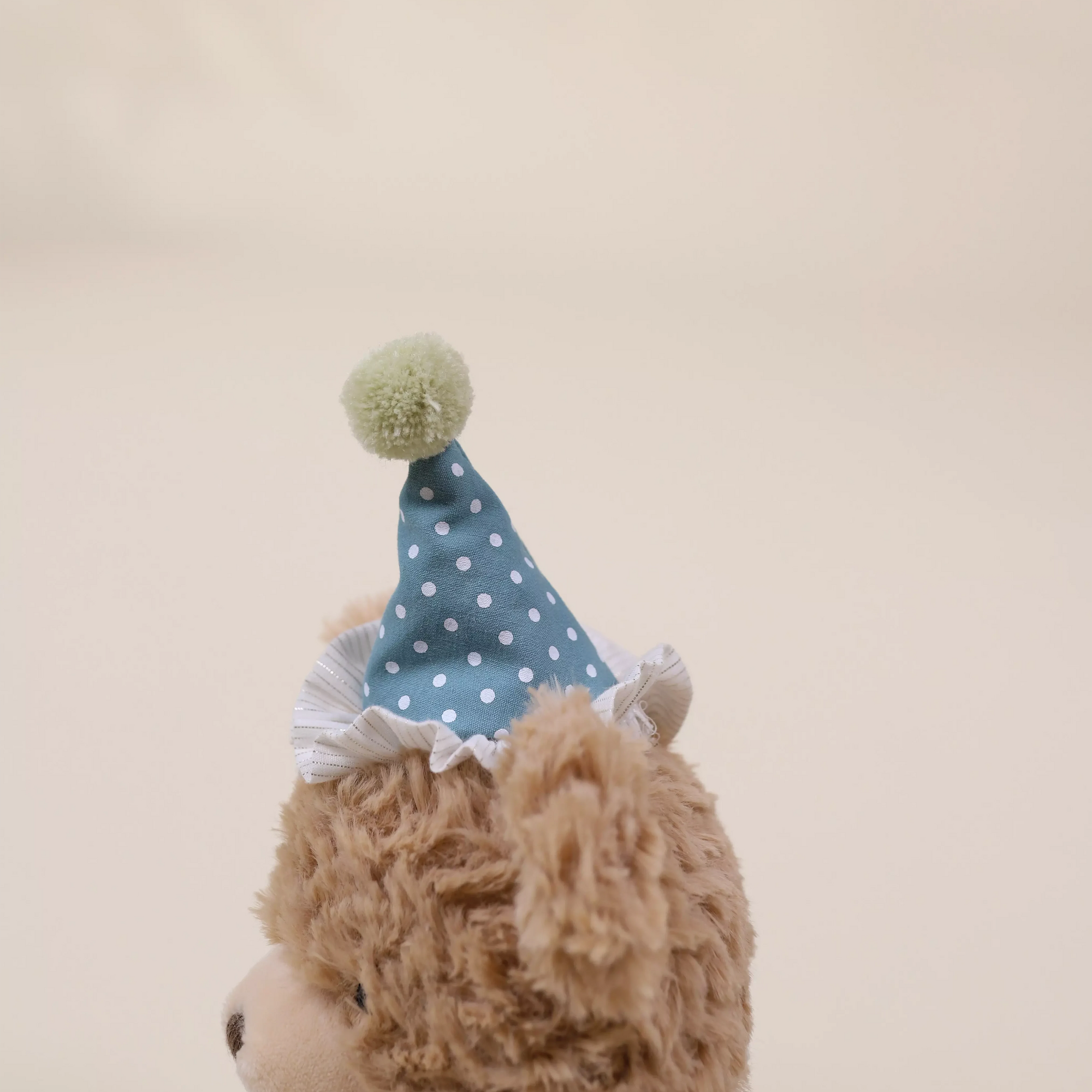 Teddy bear wearing a blue polka dot party hat with a green pom-pom on a beige background