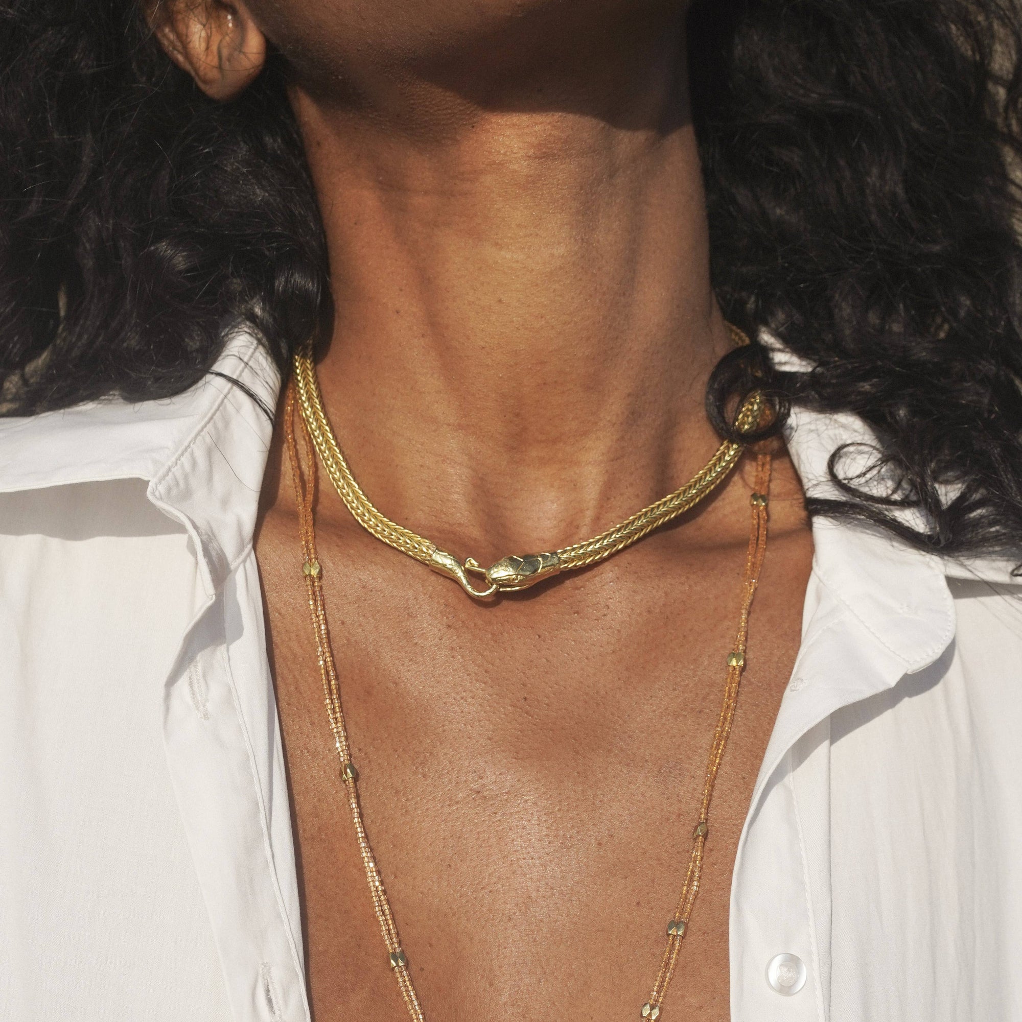 Close-up of a person wearing layered gold necklaces, including a snake choker and a longer snake-print chain, styled with a white button-down shirt.