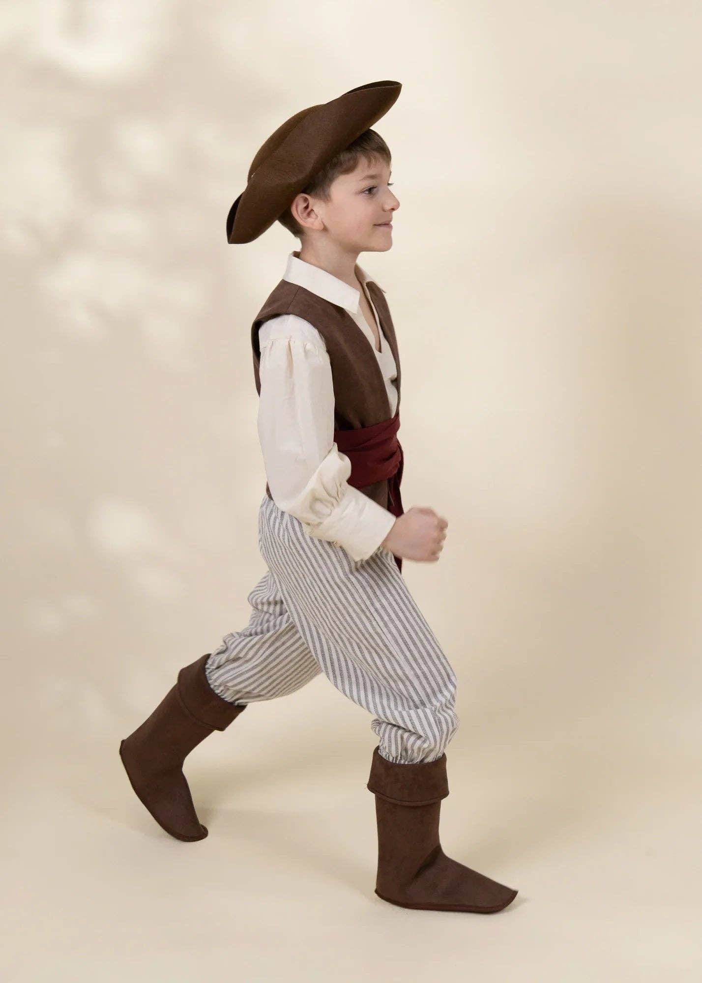A young boy modeling a pirate costume consisting of a brown hat, brown vest, white shirt, striped pants, and brown boots, set against a warm beige studio background.