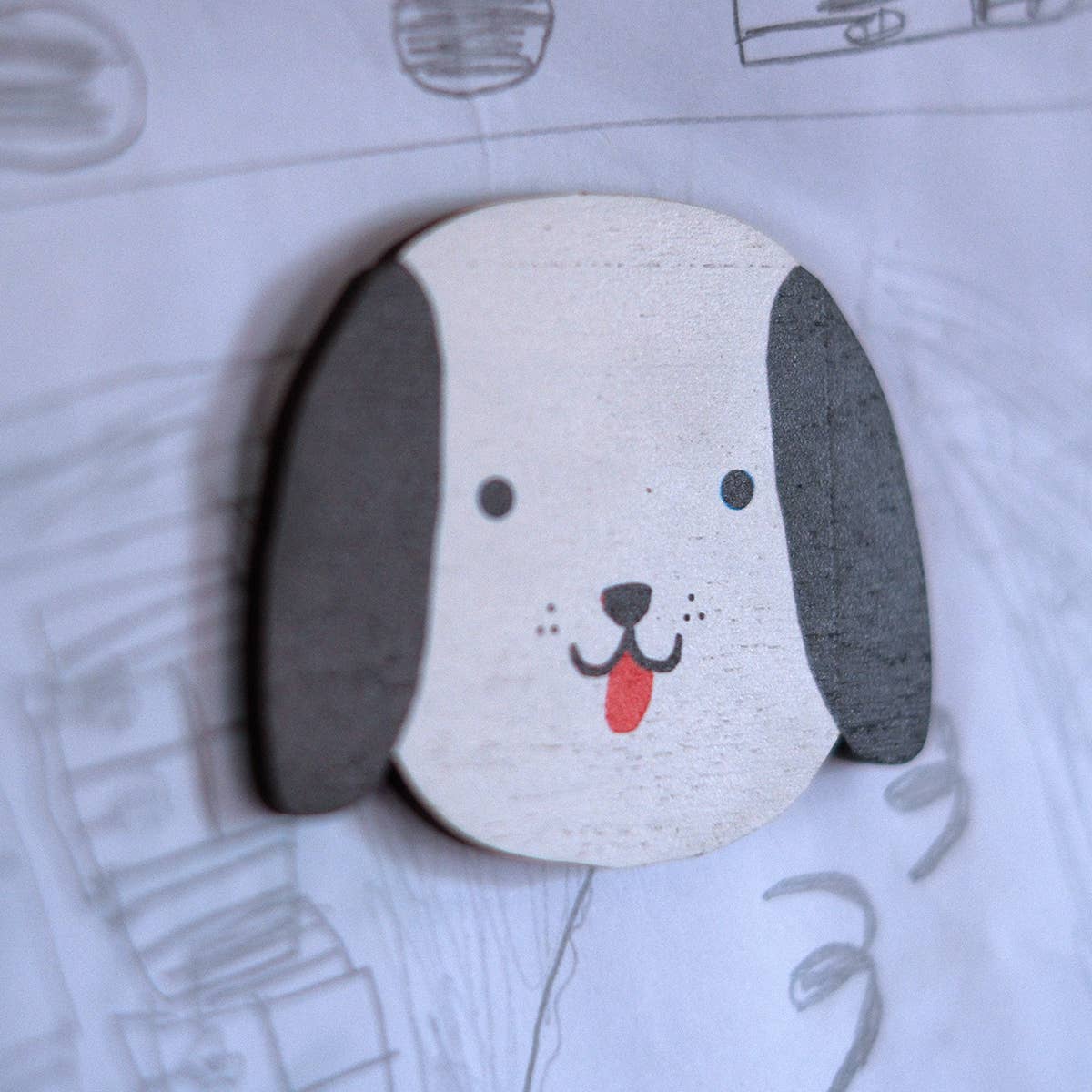 A round wooden dog head pin or badge with a white face, dark gray ears, and a red tongue, displayed on a light background with faint pencil drawings.