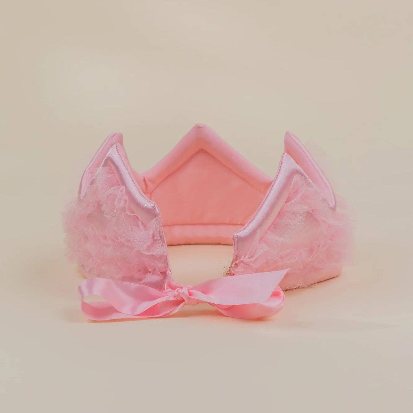 A pink plush cat-ear headband with fluffy tulle details and a matching pink satin bow, displayed on a solid light beige background.