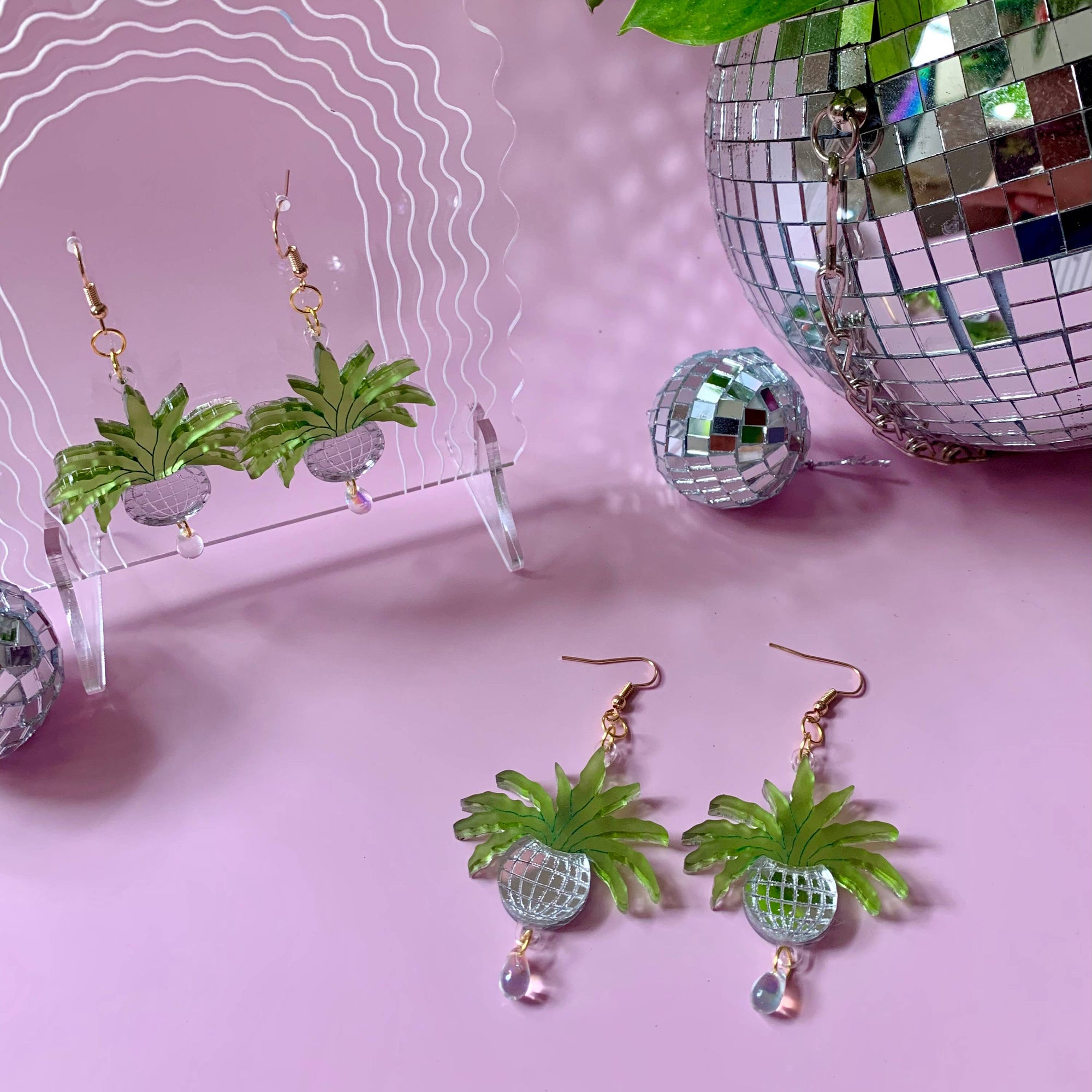Green pineapple-shaped earrings with clear gemstones on a pink background with disco ball and plant.