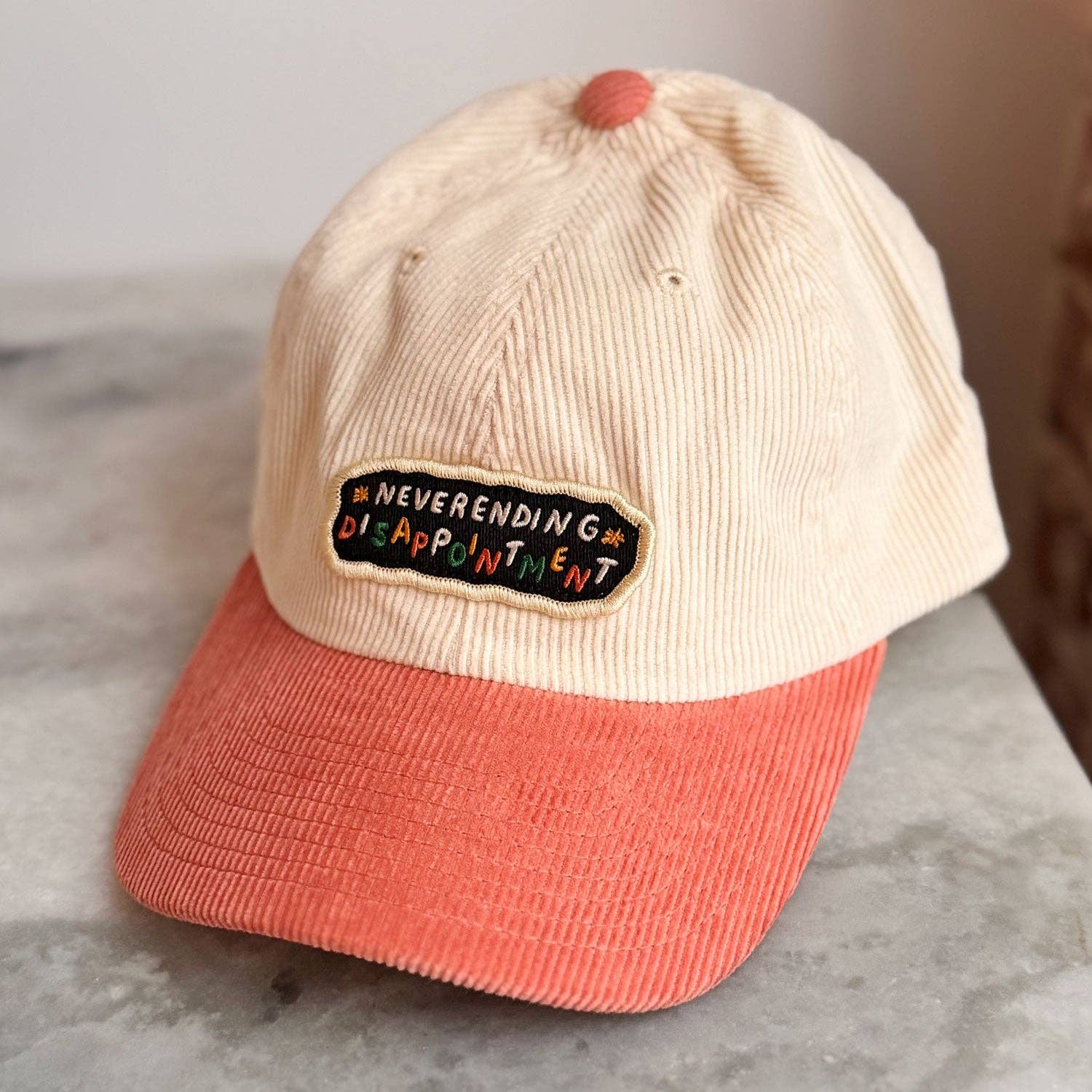 Beige and pink cap with 'Neverending Disappointment' patch on a marble surface