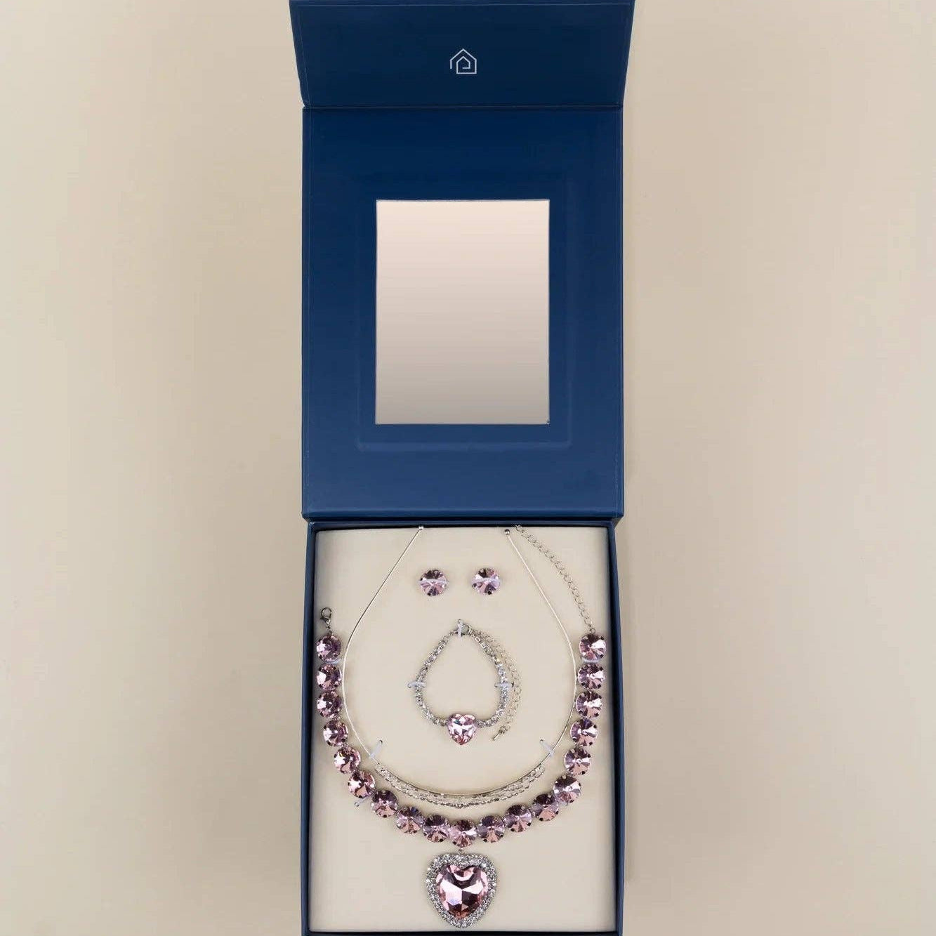 A silver-toned necklace and matching earrings set with light purple gemstones, presented in an open dark blue jewelry box with a mirror inside, set against a plain beige background.