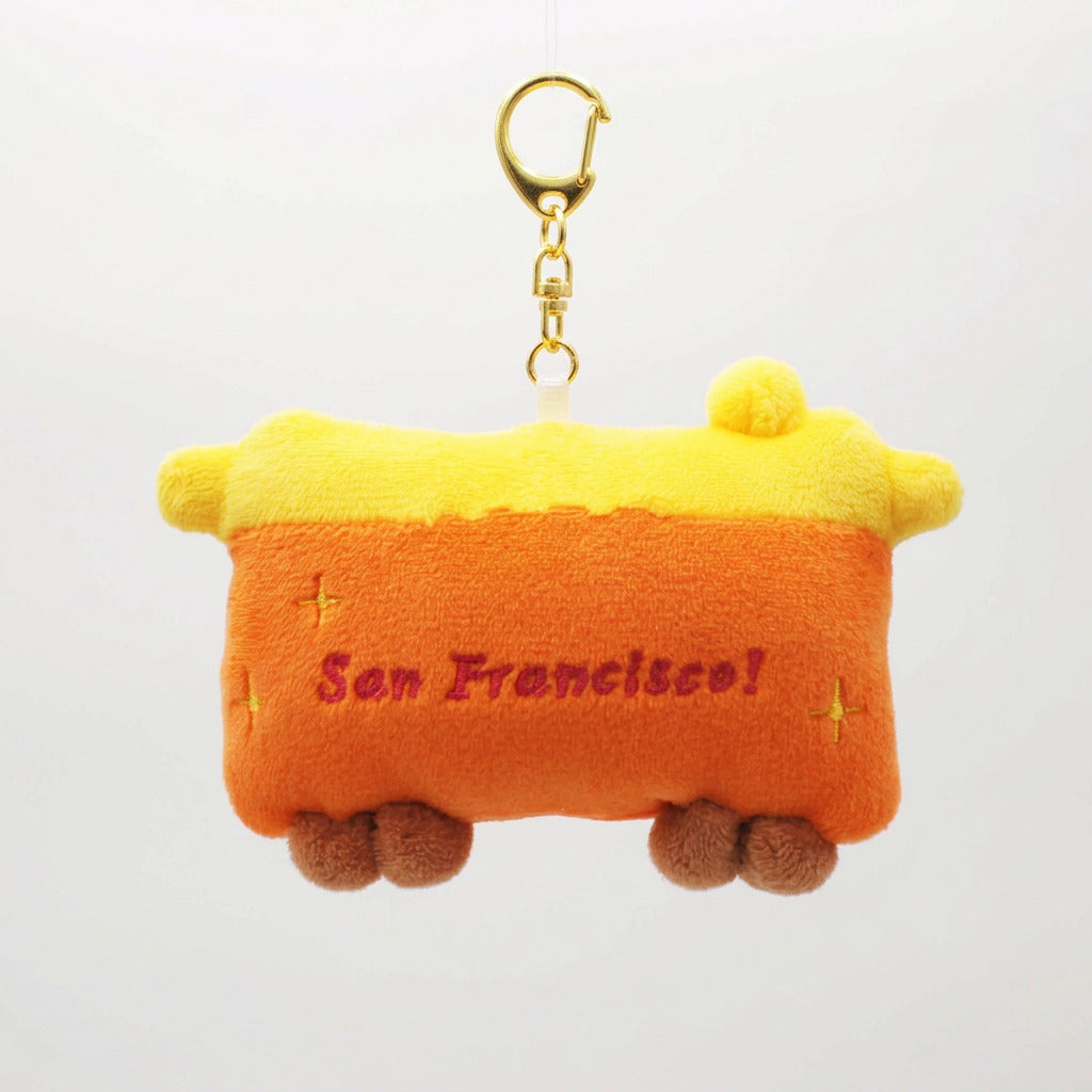 A plush San Francisco train-shaped keychain bag in orange and yellow, with 'San Francisco!' embroidered in red on the front, attached to a black strap and gold clasp, shown against a white background 