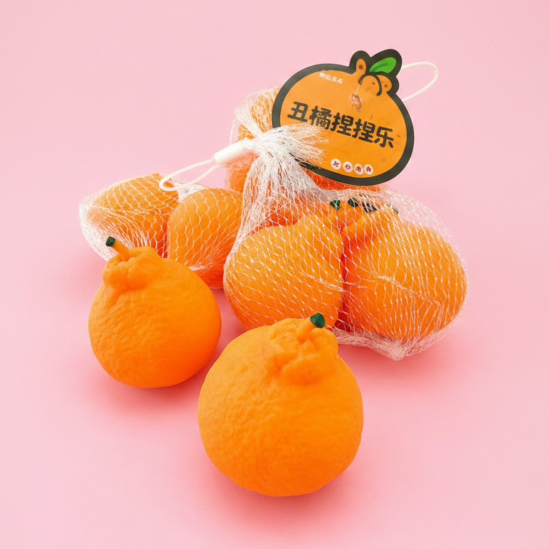 Several bright orange citrus fruits, including two loose oranges in the foreground and a mesh bag of oranges with a cartoon tag, arranged on a solid pink background.