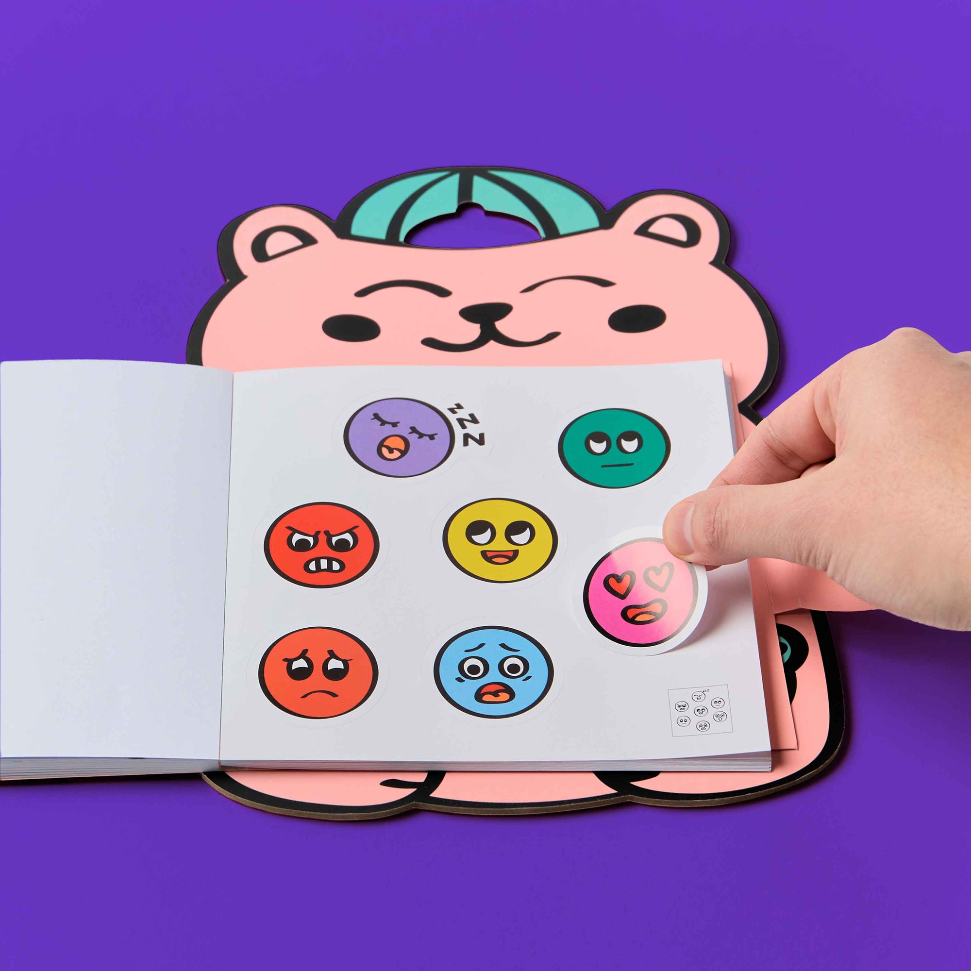 Colorful emoji stickers on a book held by a hand with a pink bear-shaped object in the background.