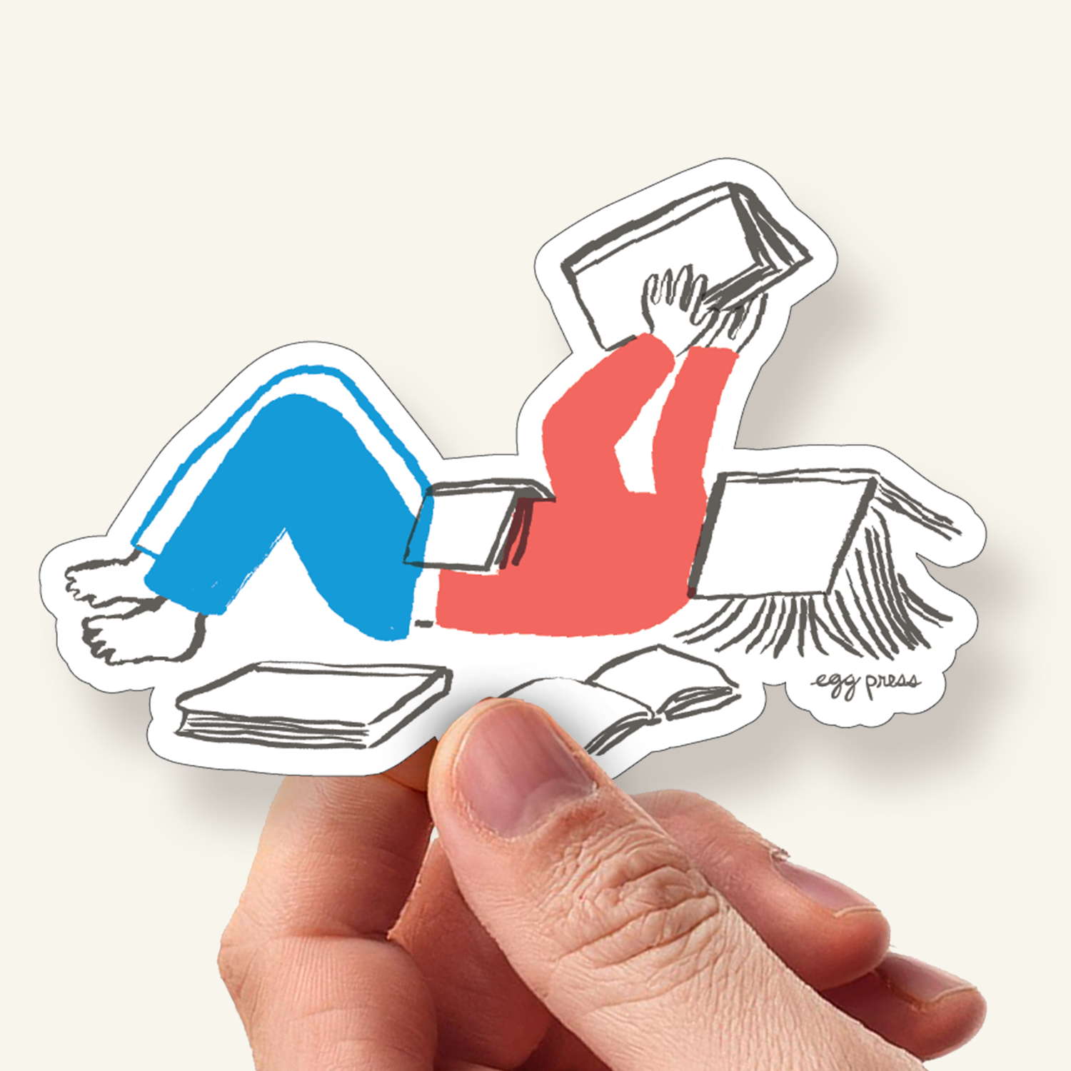 Sticker of a person lying down reading books held by a hand, with a hand holding the sticker.