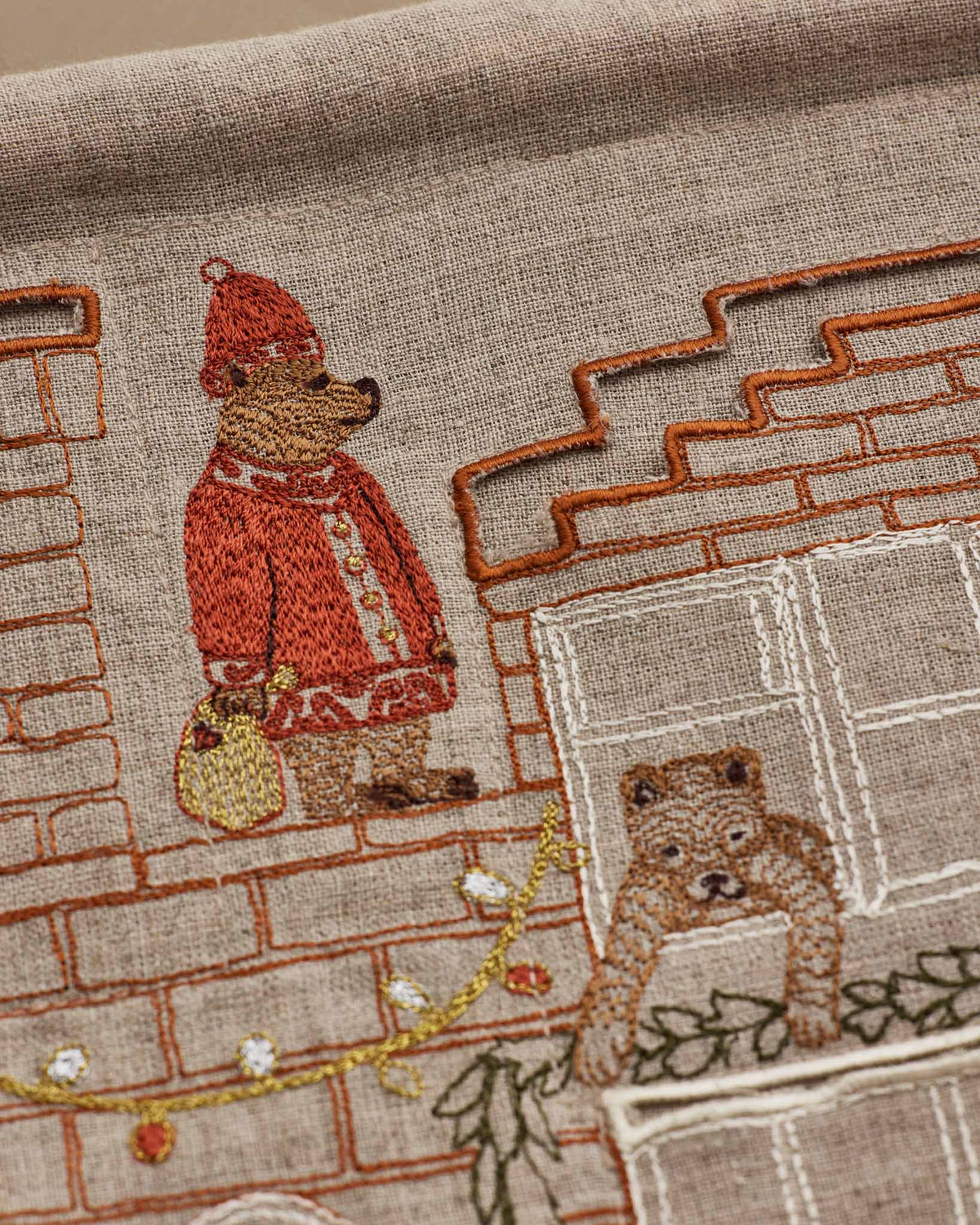 Close-up of embroidered textile featuring two teddy bears in front of a brick house. One bear wears a red coat and hat and holds a yellow bag, while the other sits by a window.