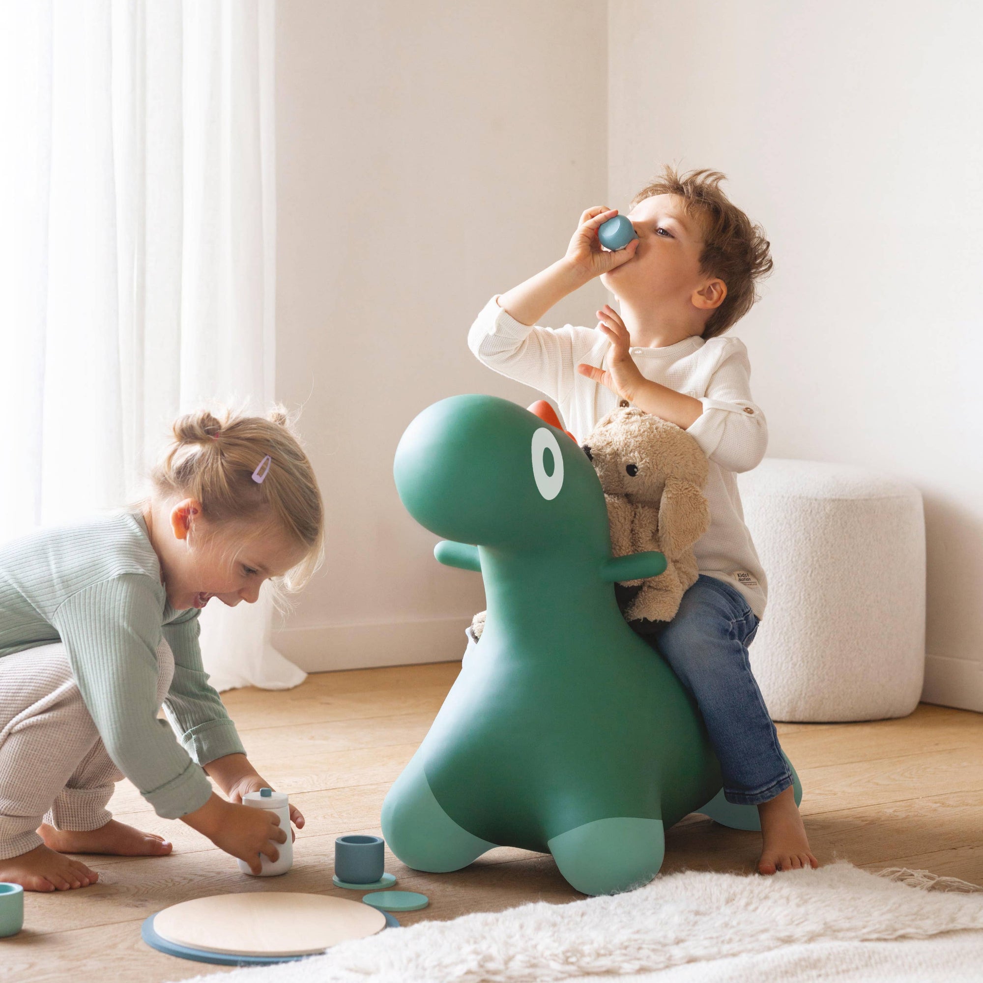 Two children playing with a green dinosaur-shaped toy in a room with light-colored walls and wooden floor.