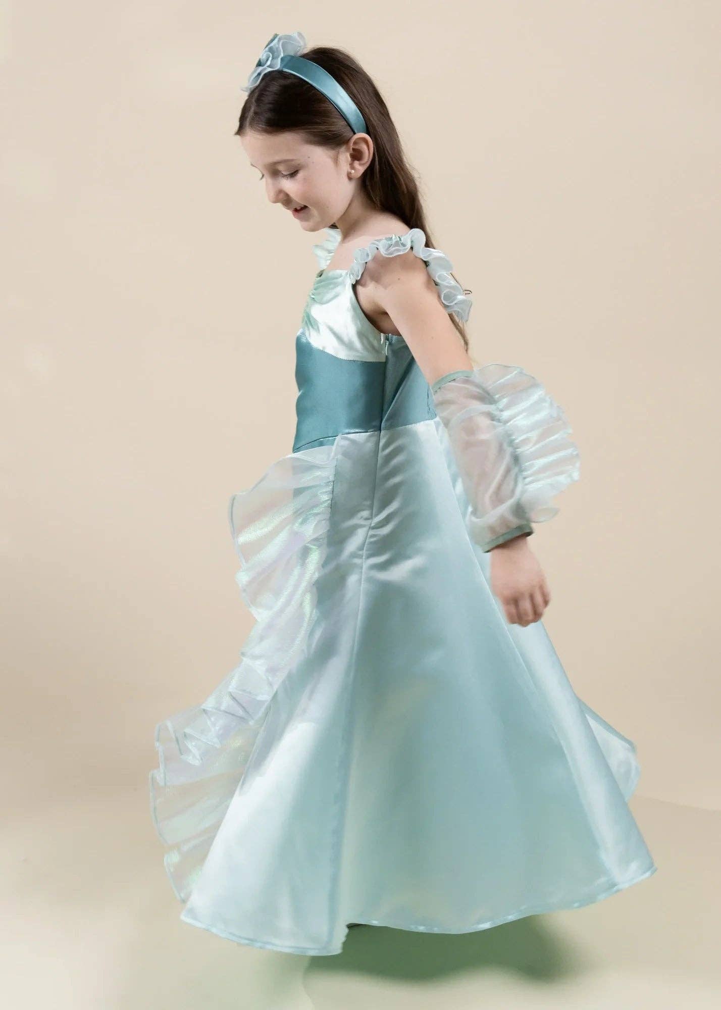 A young girl wearing a light blue princess costume dress with sheer ruffled sleeves, a matching headband, and a tiara, posing against a solid beige background.