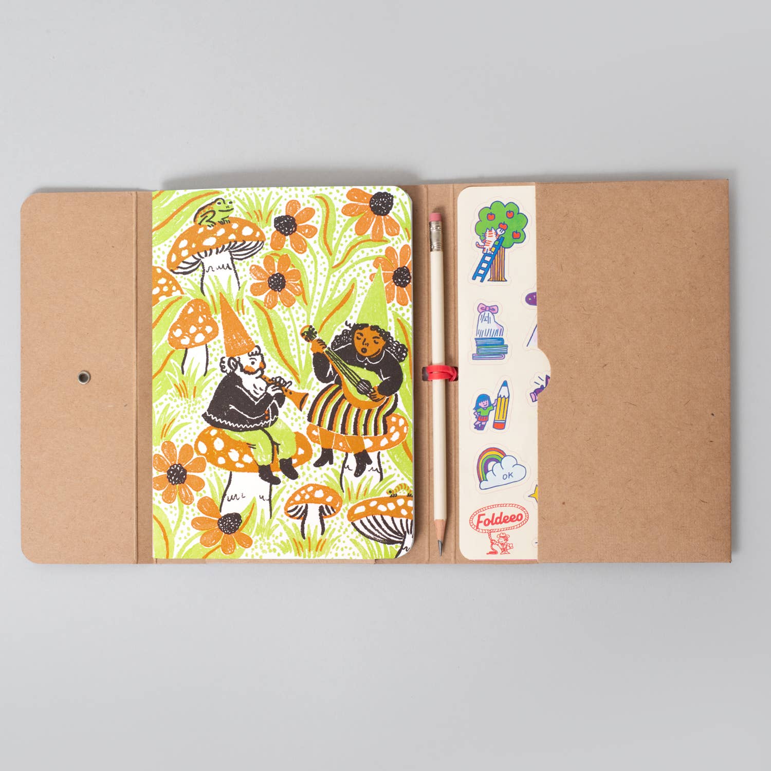 A kraft paper notebook cover opened flat on a light gray background, showing a colorful illustrated floral and gnome pattern on the left page and a pocket on the right holding a pencil and small stickers. The brand name Foldface is visible on a sticker.