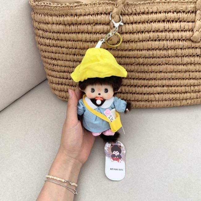 Plush keychain of a character with a yellow hat and blue outfit held by a hand against a woven basket background.