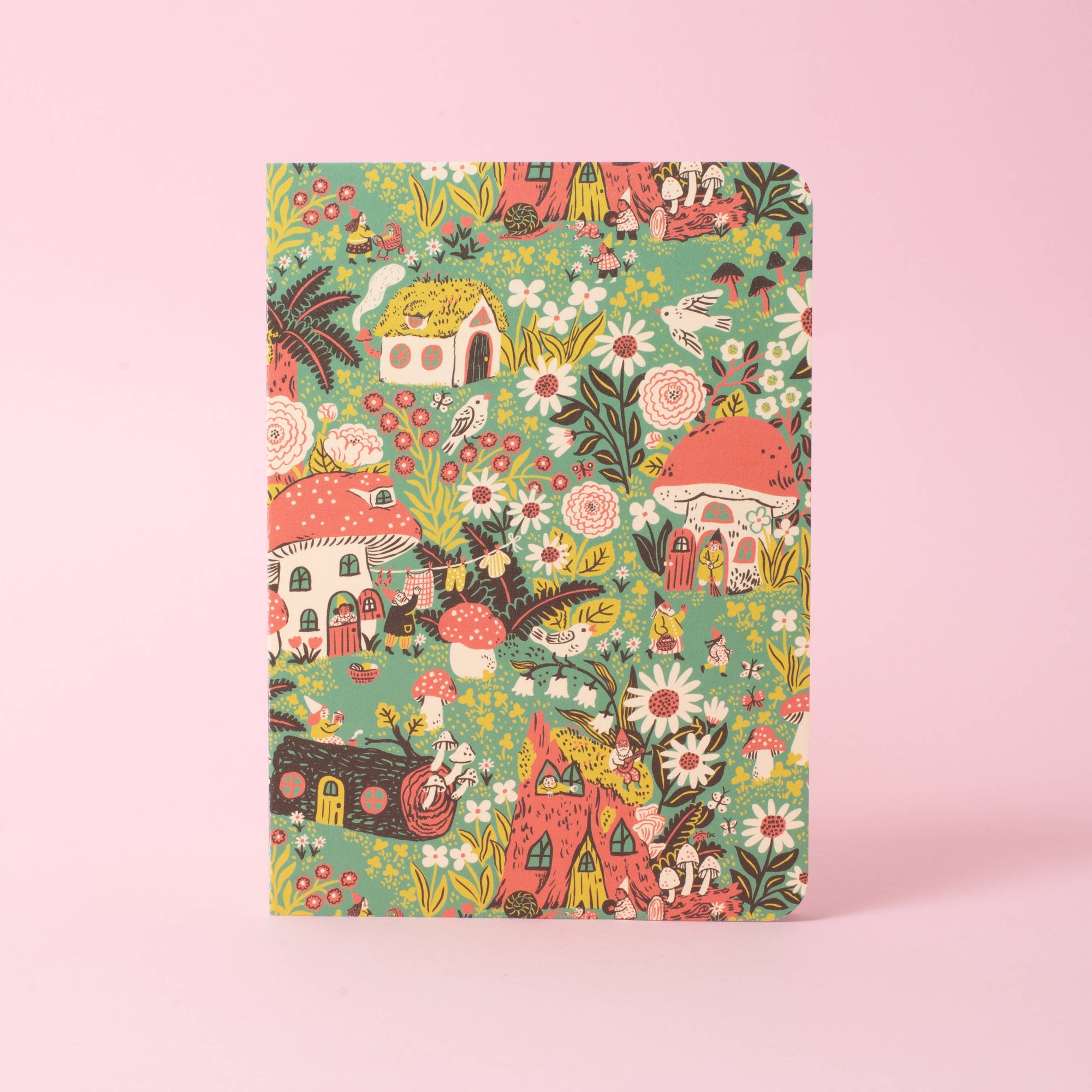 A hardcover notebook with a whimsical, colorful forest illustration featuring mushrooms, houses, flowers, and small creatures on a mint green background, set against a solid pink backdrop.