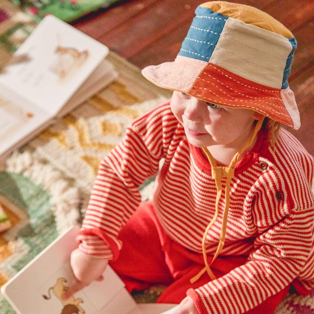 Child wearing a colorful hat and red outfit, sitting on a patterned rug with a book.