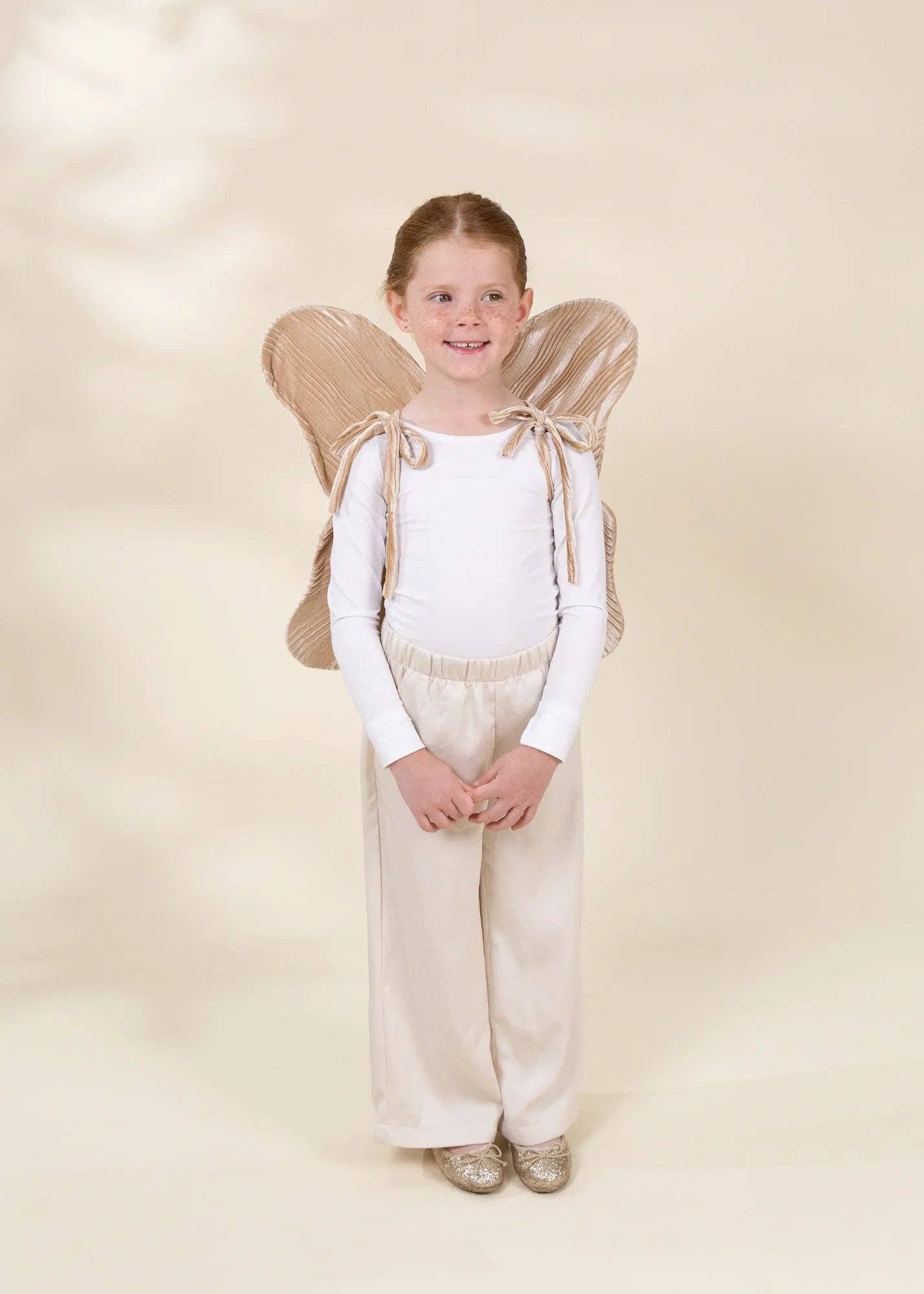 A young girl stands against a warm beige studio background wearing a white long-sleeve top, cream wide-leg pants, and a beige angel costume with attached wings.