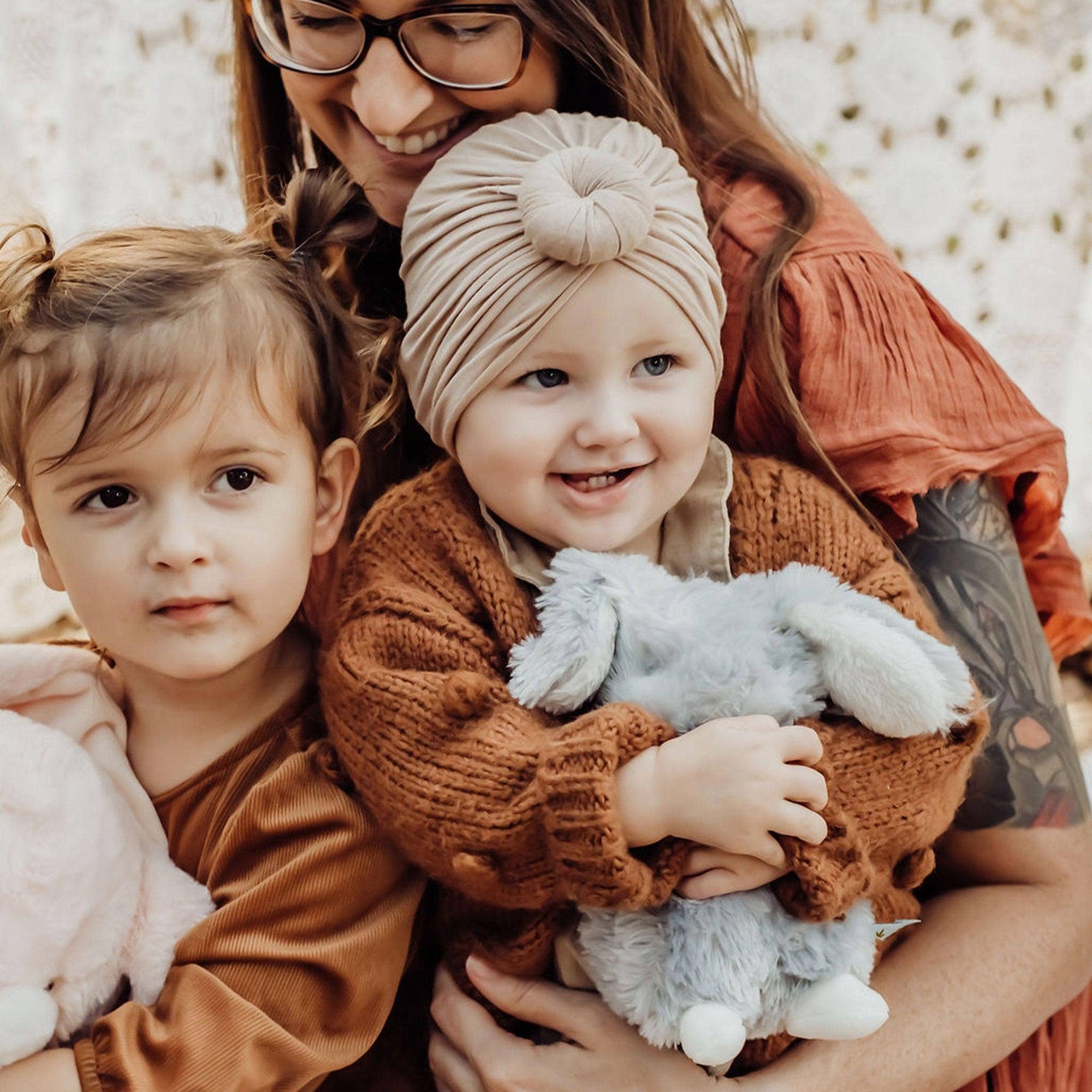 Woman holding two children, one of whom is holding a stuffed bunny, all wearing brown and beige clothing.