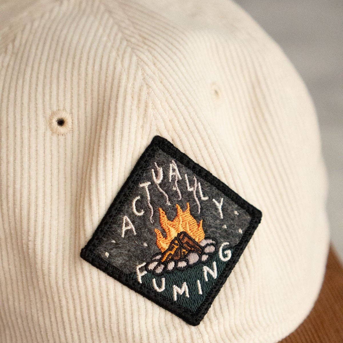 Beige cap with a patch featuring a campfire design and text.
