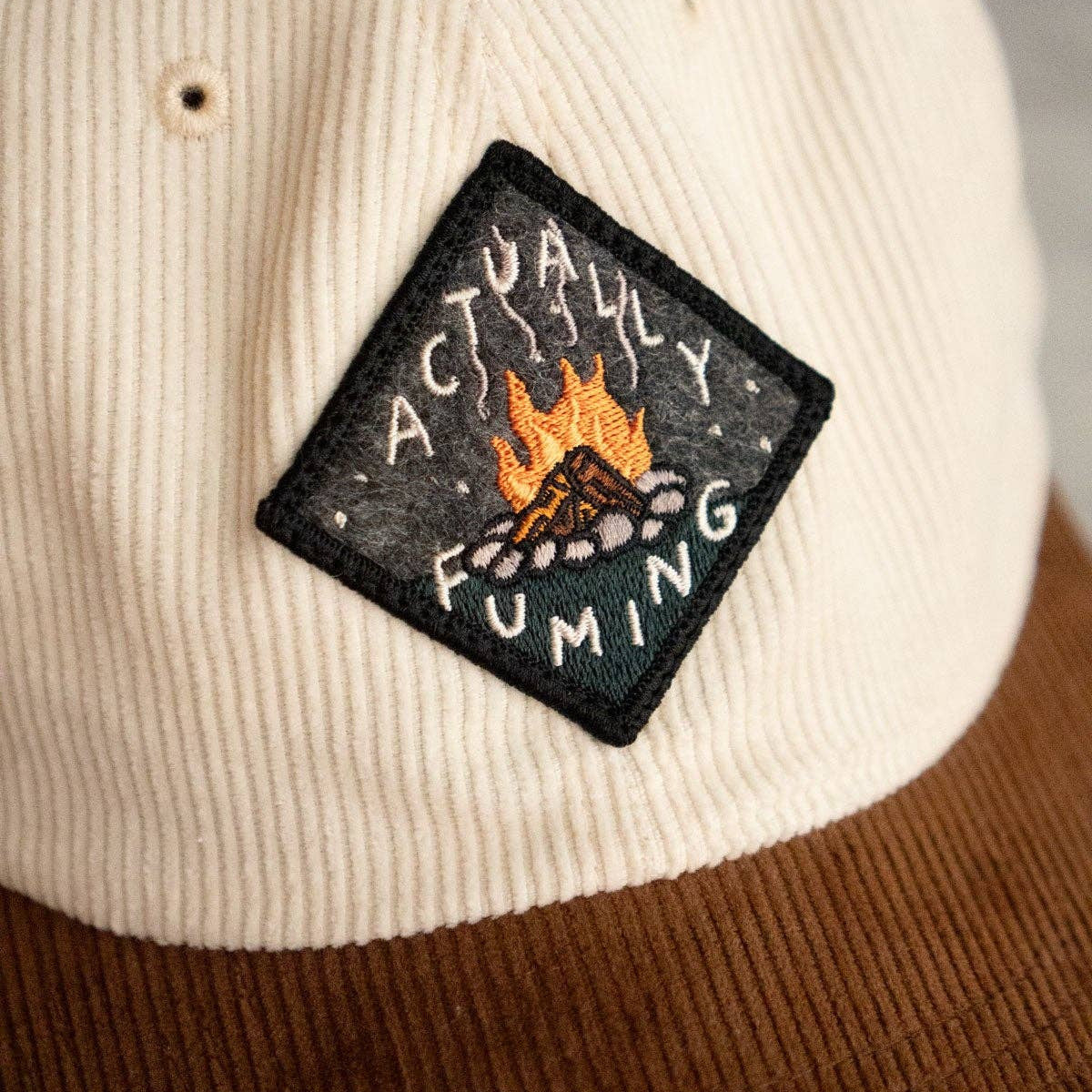 Beige cap with a patch featuring a campfire design and text.