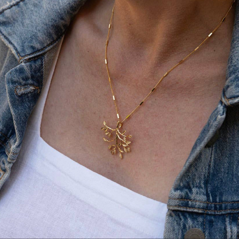 Person wearing a gold necklace with a leaf pendant, dressed in a denim jacket and white shirt.