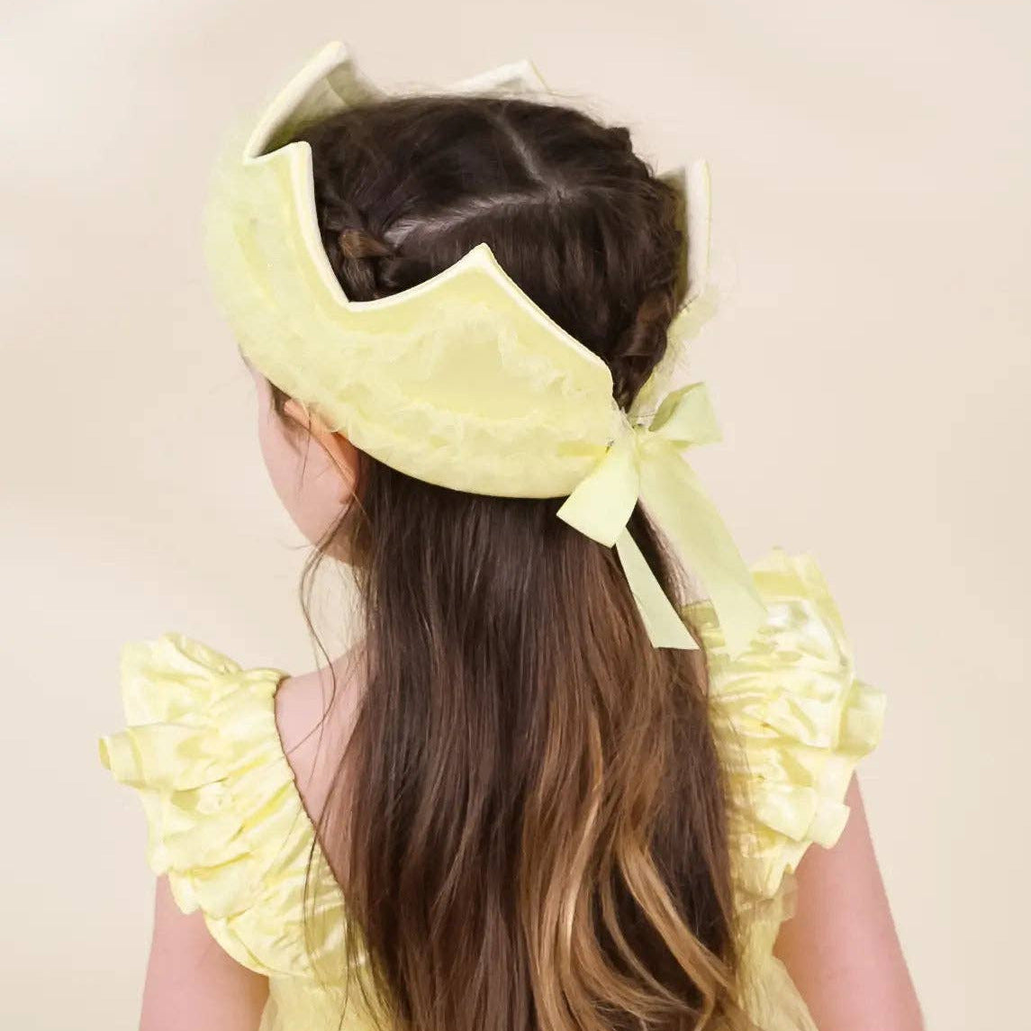A young girl viewed from behind wearing a pale yellow ruffled bonnet with a ribbon bow and a matching pale yellow dress against a plain cream background.