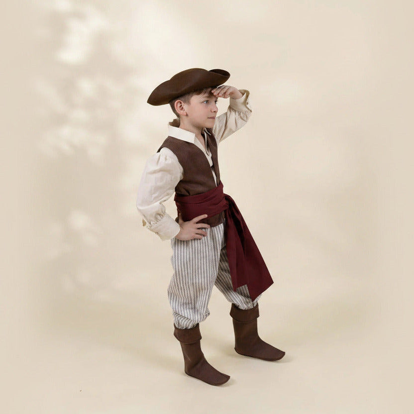 A young boy modeling a pirate costume against a warm beige background with soft leaf shadows. The costume includes a brown hat, brown vest, white and brown pinstriped pants, a dark red sash, and brown boots.